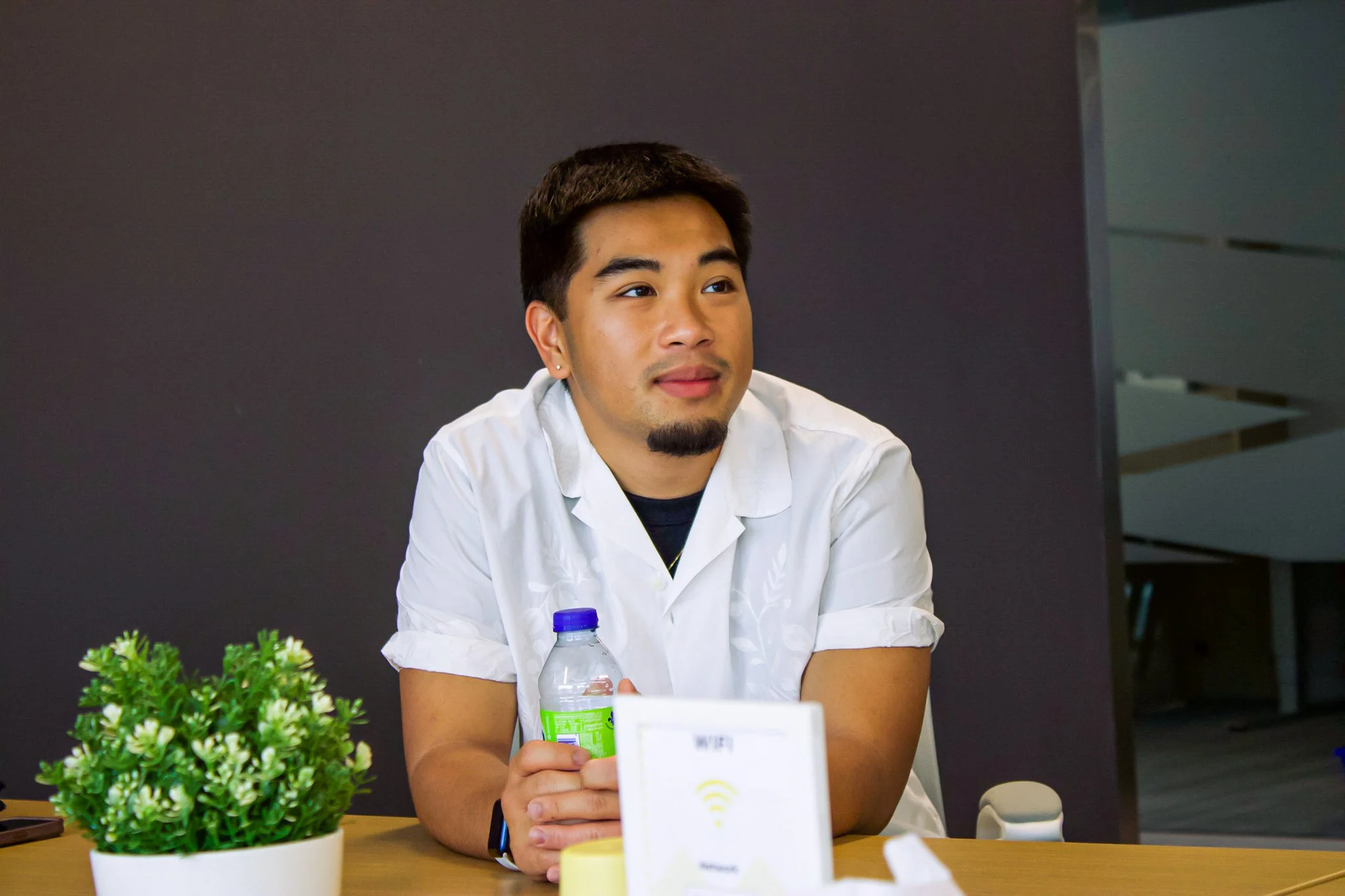 Young man sitting at a table with a plant, holding a water bottle, and looking to the side in a modern office or meeting room.