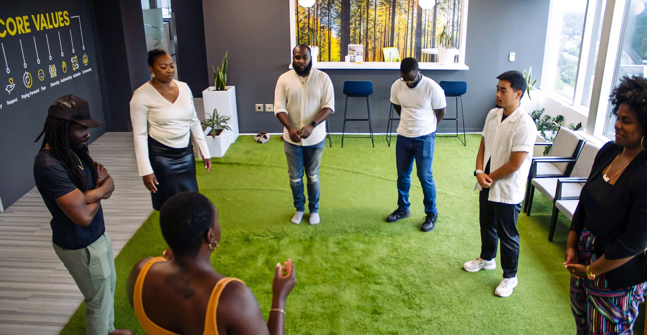 A diverse group of seven people standing in a circle in an office with green carpet and large windows, engaging in a group activity or discussion.