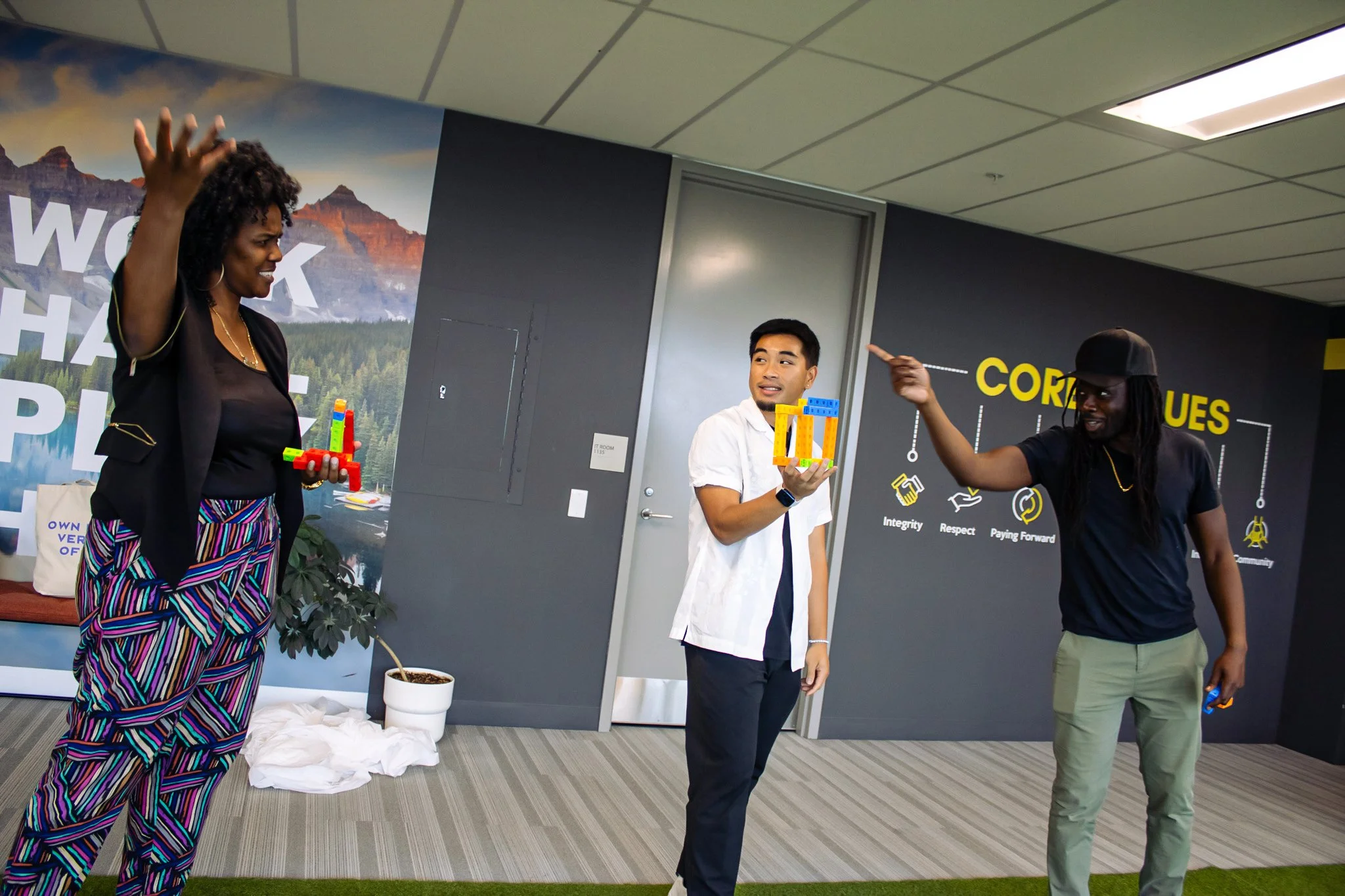 Three people standing in a modern office, engaging with colorful building blocks, with a world map and motivational words on the wall in the background.