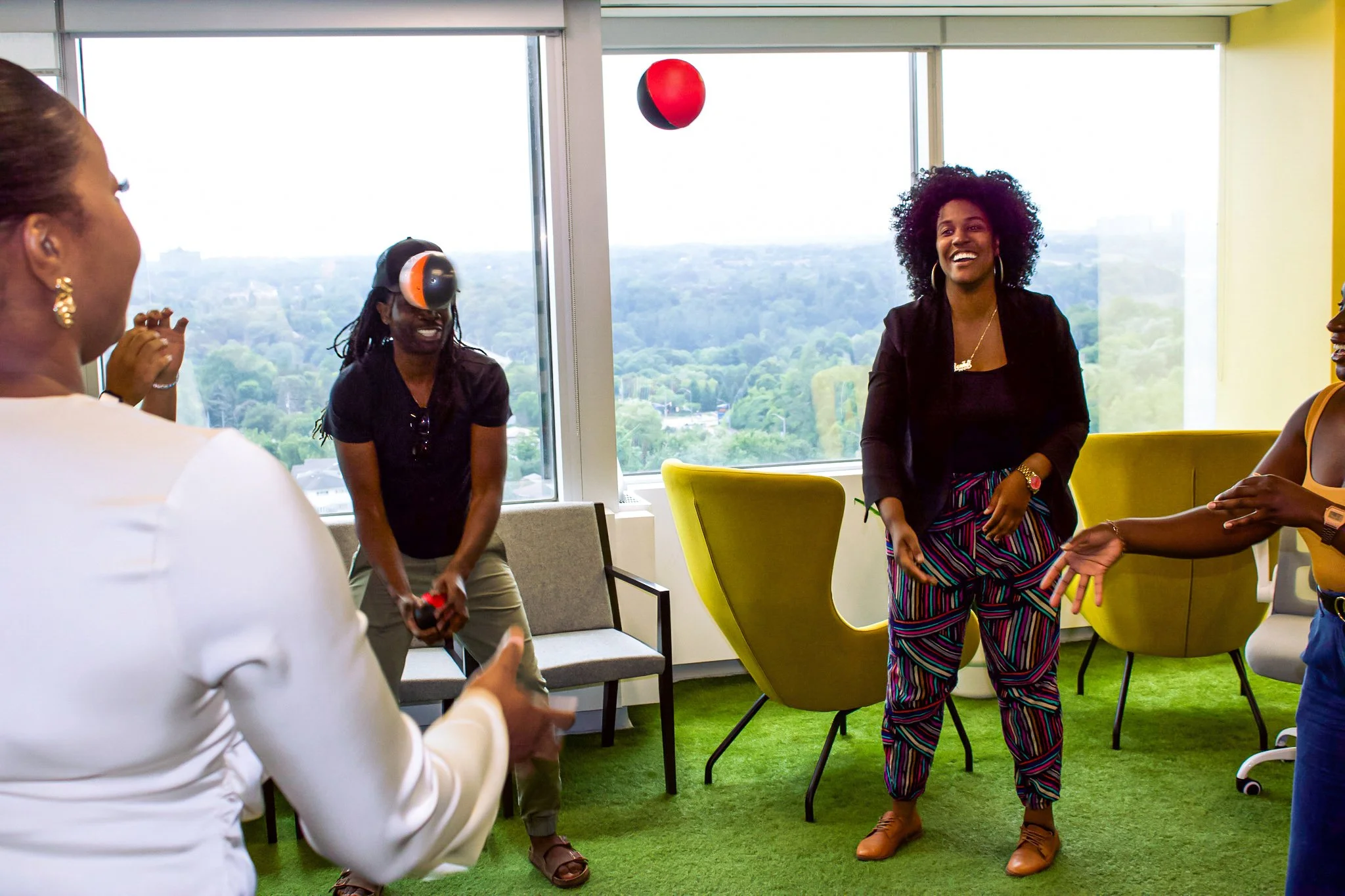 A group of women playing a VR game together in a bright office lounge with green carpet, modern chairs, and large windows showing an outdoor view.