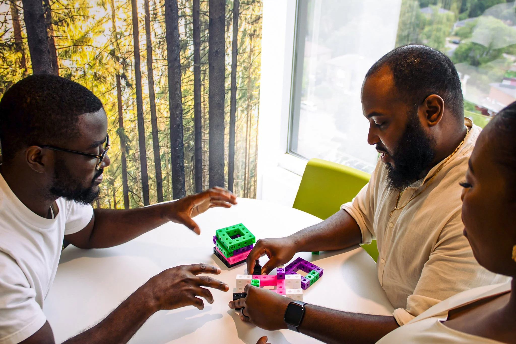 Three people playing with colorful building blocks at a white table near a window, with a forest scene visible outside.