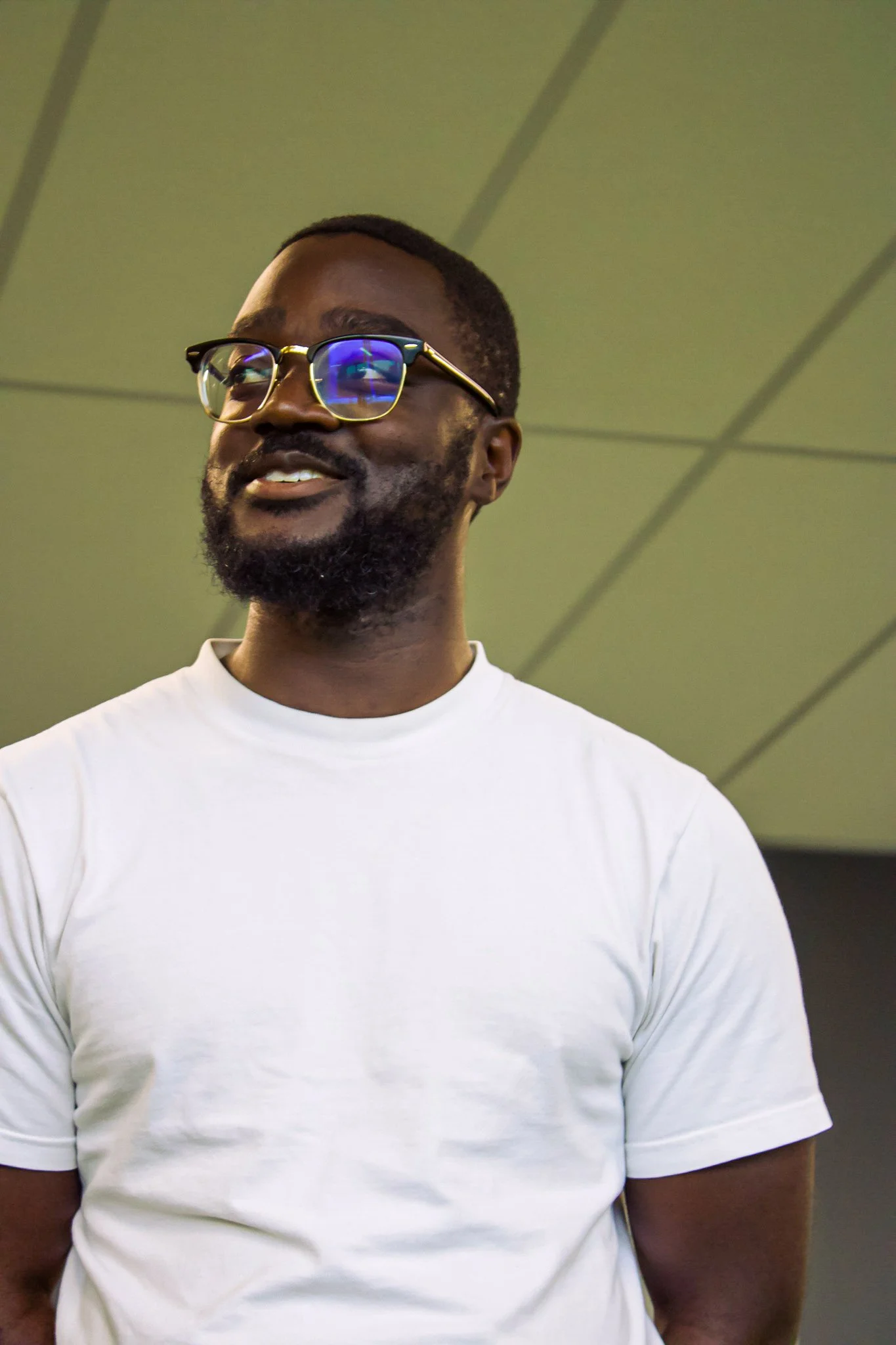 A smiling man with glasses and a beard, wearing a white t-shirt, stands indoors against a gray ceiling with lines.