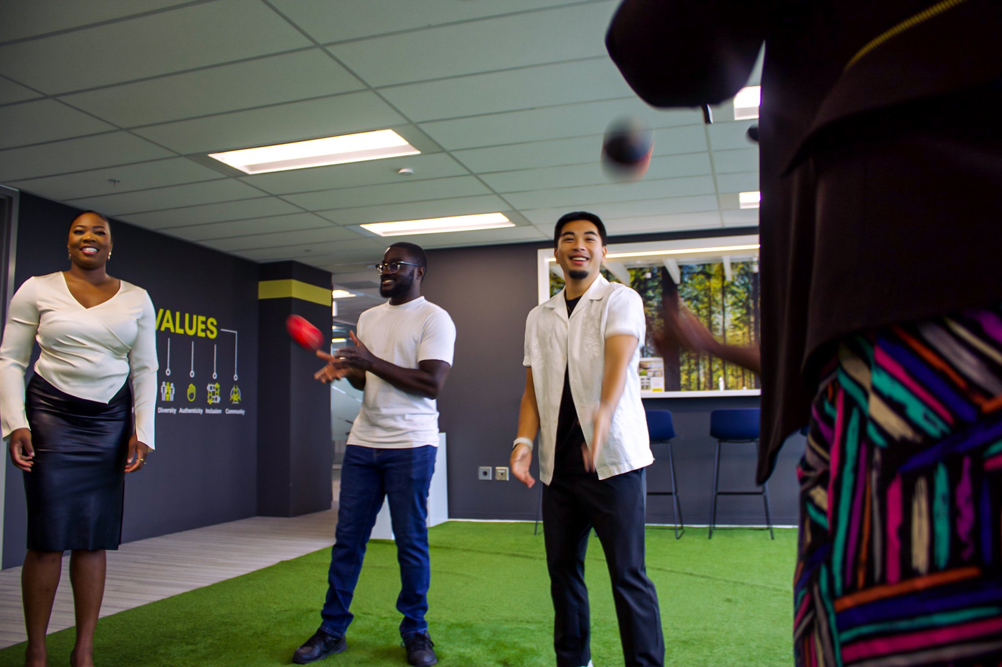 Four diverse people standing and smiling in an office setting. One woman on the left wearing a white blouse and black skirt, two men in the middle wearing white shirts, one with glasses, and another with a black shirt underneath, and a person on the right partially seen wearing a brown shirt with colorful stripes. They seem to be playing with red and black balls. The office has a modern design with a mural of a forest outside the window and a sign on the wall that says 'VALUES' with icons representing Diversity, Authenticity, Inclusion, and Community.