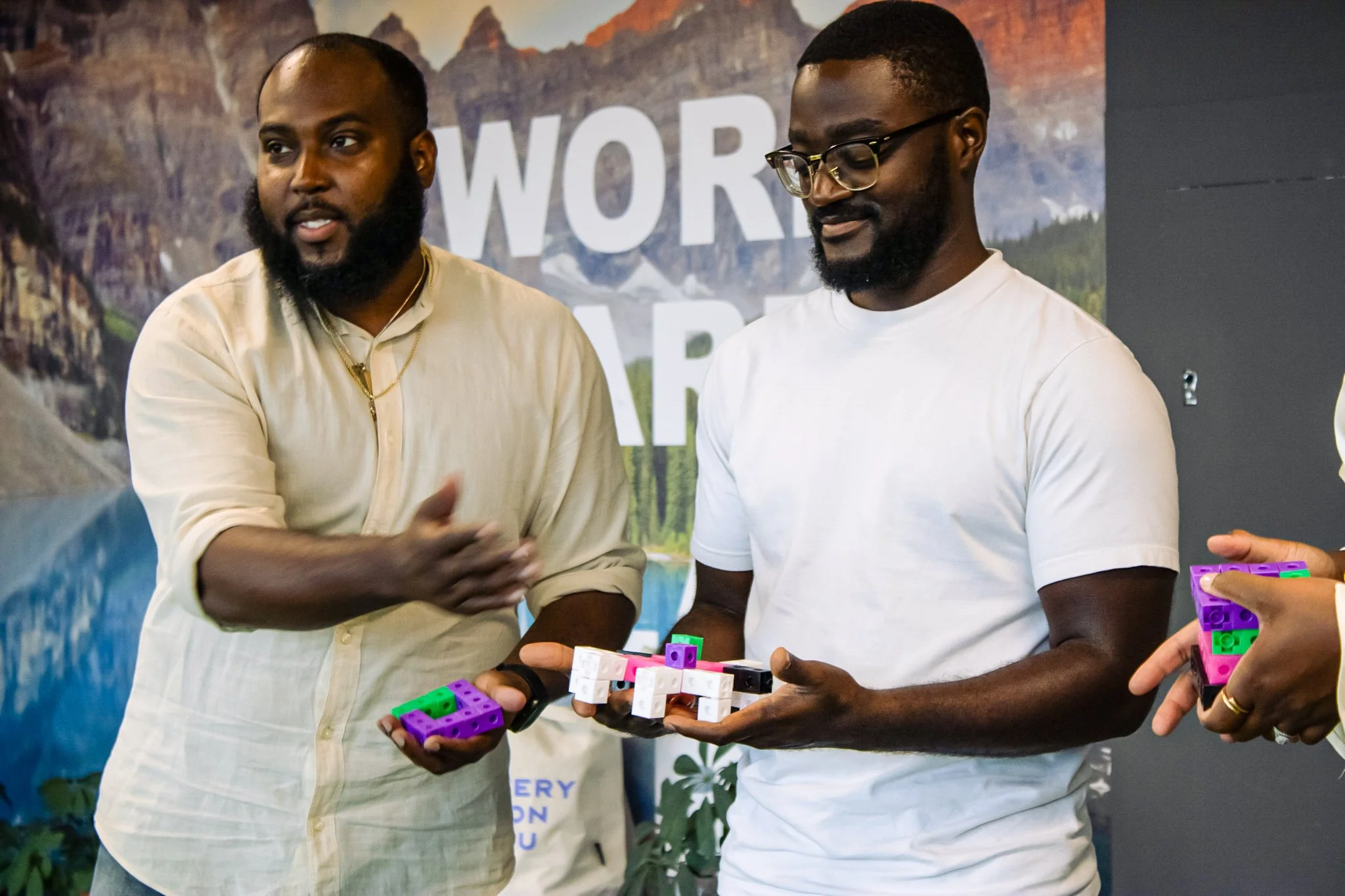 Two men standing indoors holding colorful robotic building blocks, with a mountain and lake backdrop behind them. One man is speaking, and the other is looking down at the blocks.