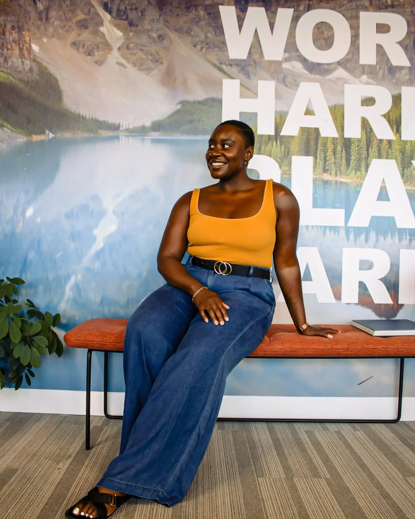 A woman sitting on a bench in front of a mural of a mountain lake, smiling and looking to the side, wearing a yellow sleeveless top, wide-leg blue jeans, and black sandals.