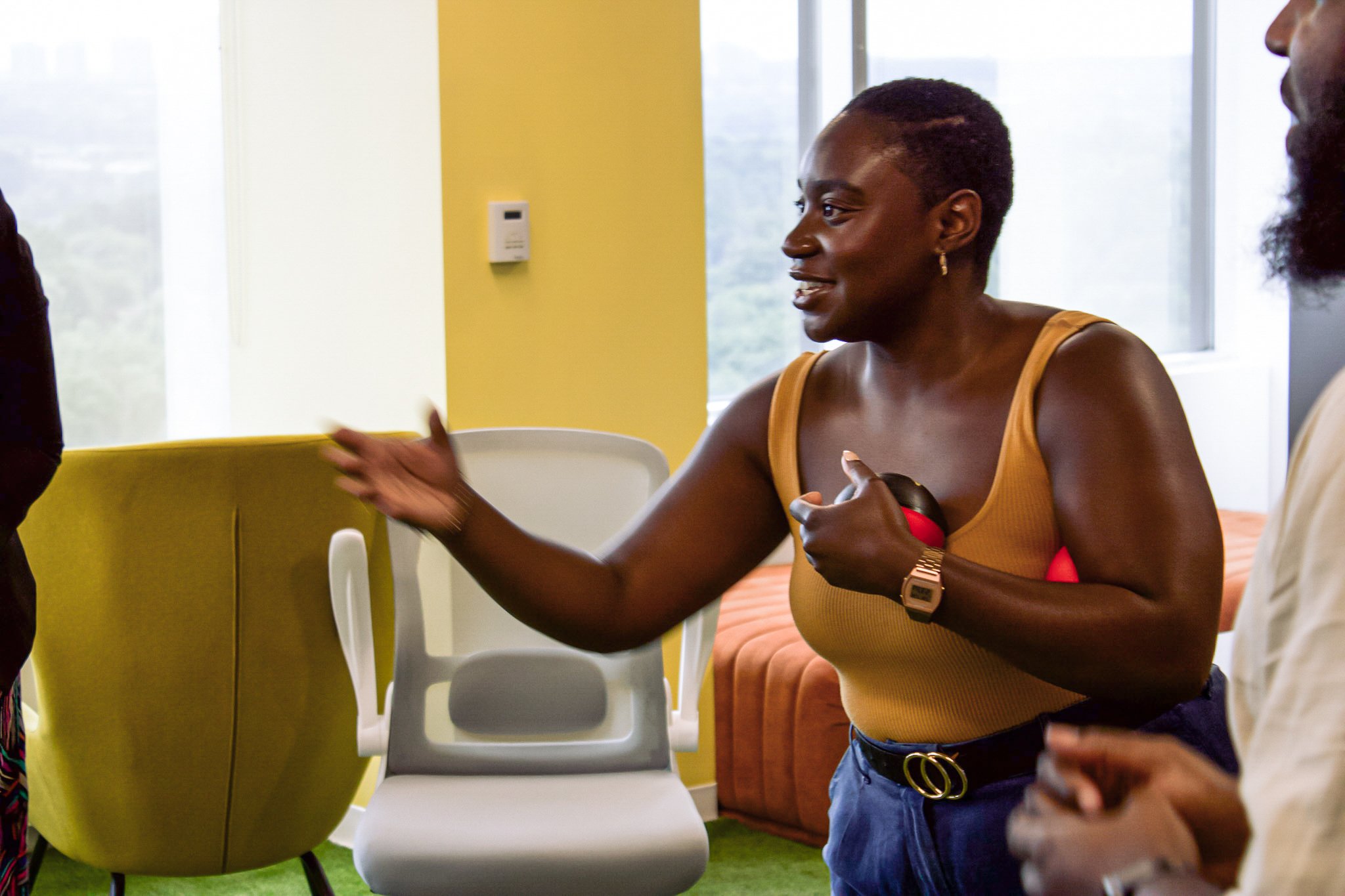 A woman in a mustard-colored sleeveless top gestures while holding a stress ball in a brightly lit room. She is wearing a watch and has a short hairstyle. There are chairs and a window in the background.