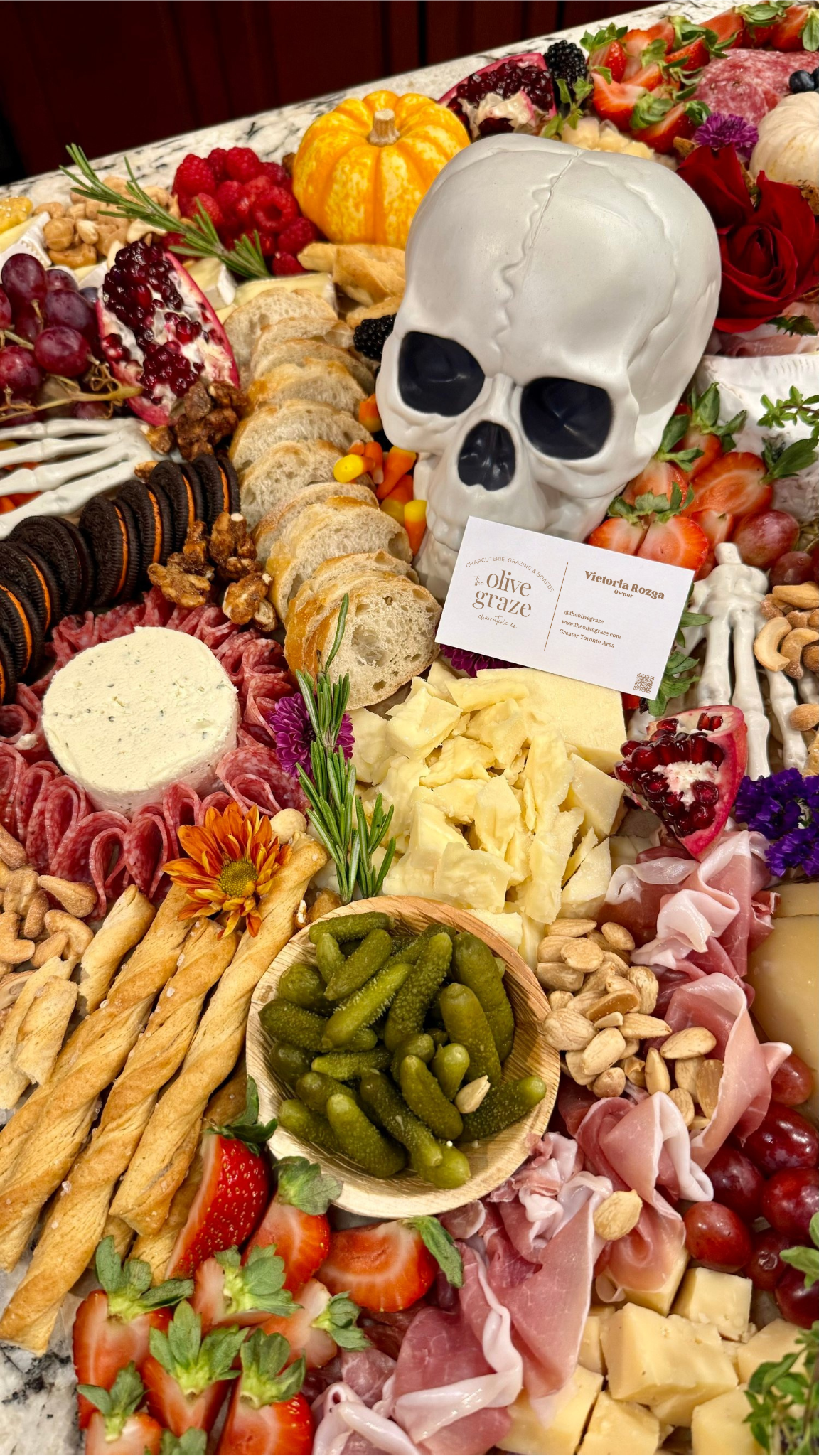 A platter of assorted food items including cheese, strawberries, grapes, blueberries, blackberries, a small pumpkin, a white skull decoration, crackers, nuts, pickles, various slices of meats, and garnishes like herbs and flowers.