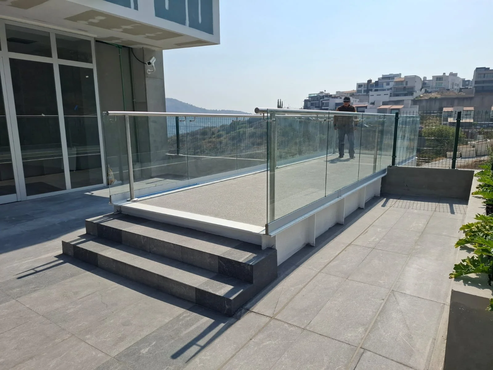 Terraza con barandal de vidrio en un edificio moderno, con escaleras de piedra y vista a zonas residenciales y montañas en el fondo.