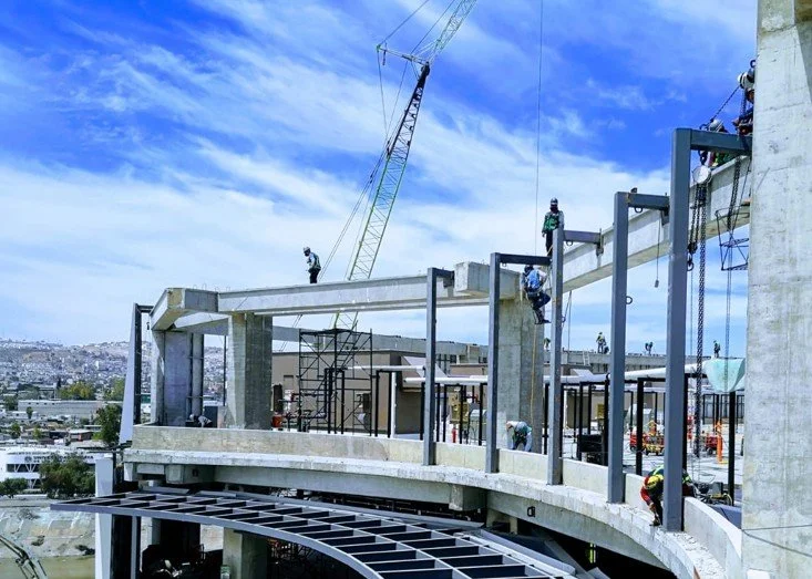 Trabajadores en construcción en un edificio en desarrollo con grúa y estructura de concreto, cielo despejado de fondo.