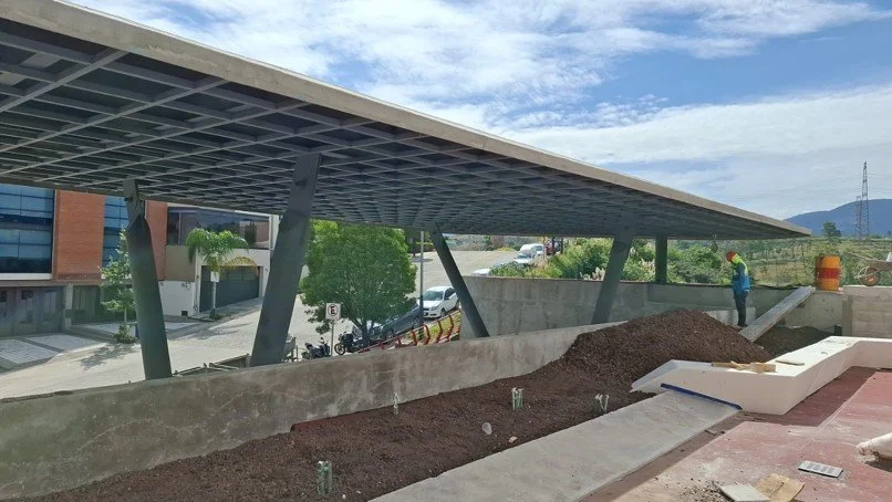 Un trabajador en un sitio de construcción cerca de una estructura moderna con un techo inclinado y columnas, con montículo de tierra y materiales de construcción en primer plano.