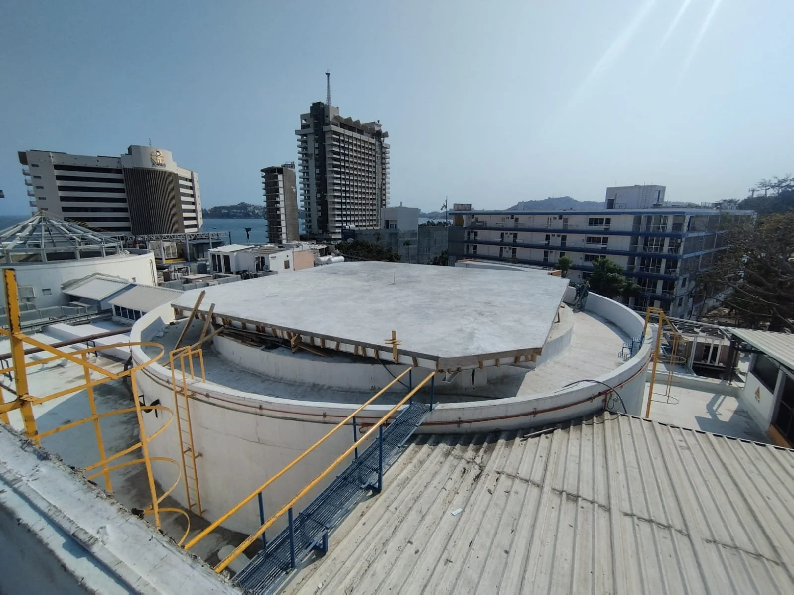 Construcción en la azotea de un edificio, en proceso de obra, con estructuras de protección y andamios, con edificios altos y una vista de un cuerpo de agua al fondo.
