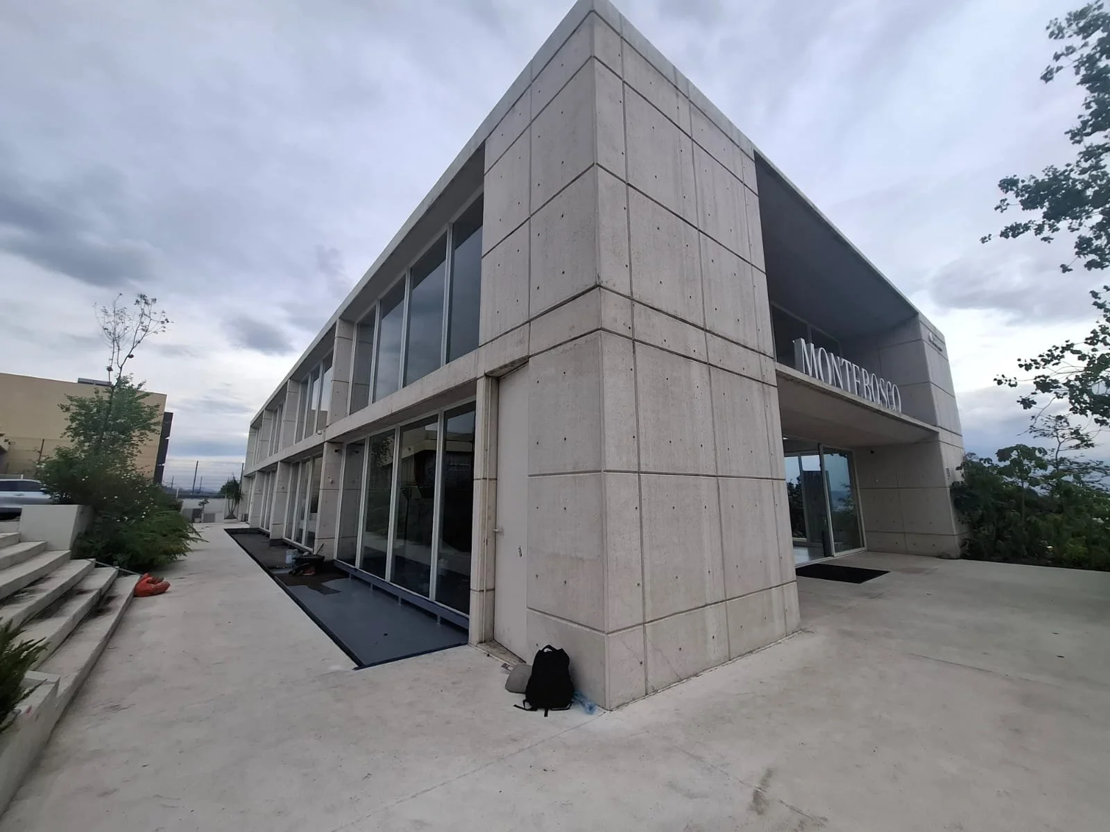Edificio moderno de dos pisos con paredes de concreto y muchas ventanas de vidrio, con el nombre 'Monterroso' visible en la fachada. El cielo está nublado y hay árboles y plantas alrededor.