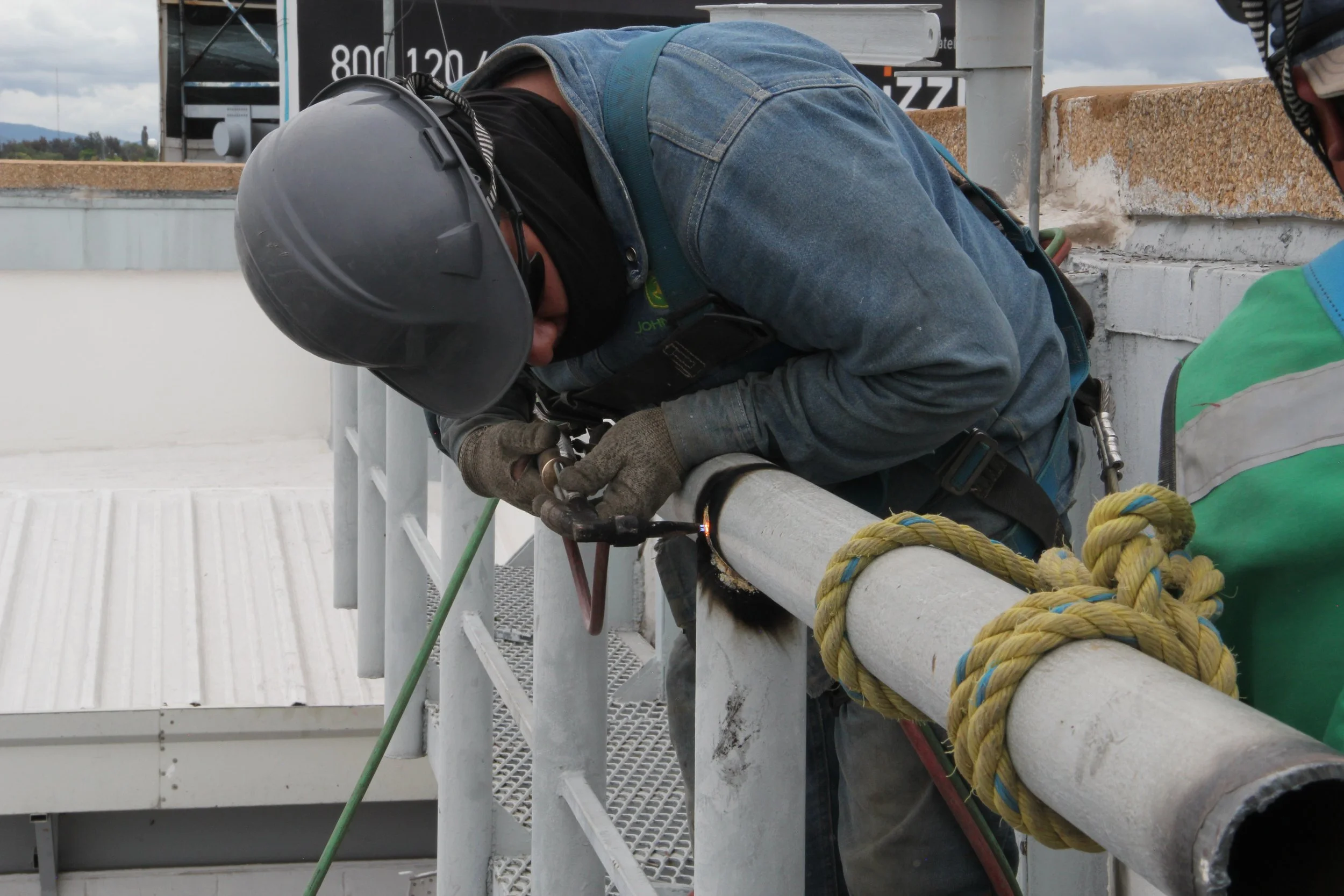 Un trabajador con casco, guantes y arnés, soldando una tubería metálica en una estructura industrial exterior, con herramientas y cuerda de seguridad.