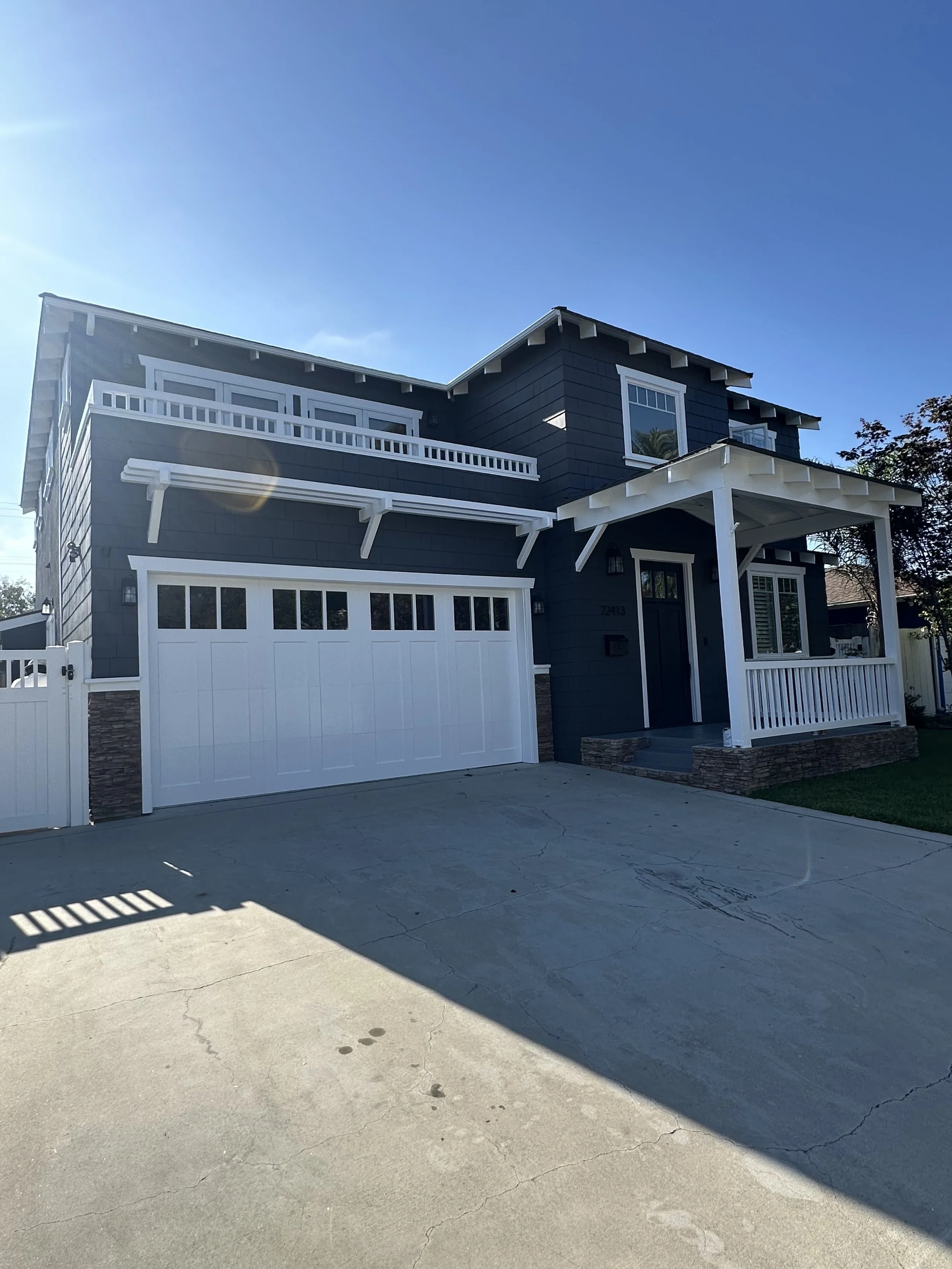 A two-story modern house with dark blue exterior, white trim, and a front porch. There is a white garage door, a driveway, and some greenery on the side. The sky is clear and sunny.