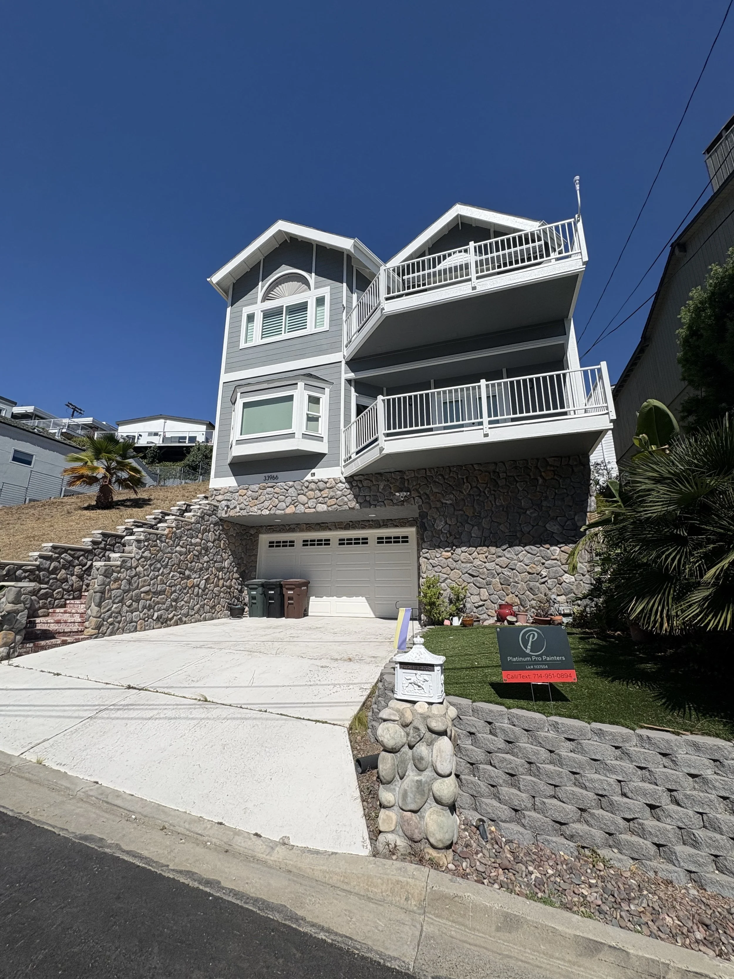 A multi-story gray and white house with large balconies and a stone foundation, situated on a sloped lot with plants and a driveway with a car parked in front.