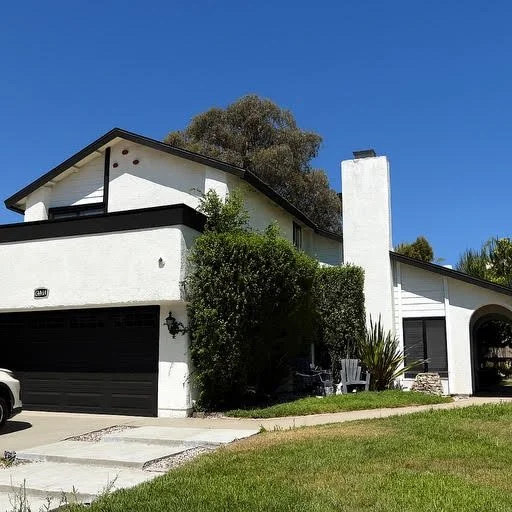 White two-story house with black accents, a black garage door, a large chimney, and lush greenery, under a bright blue sky.