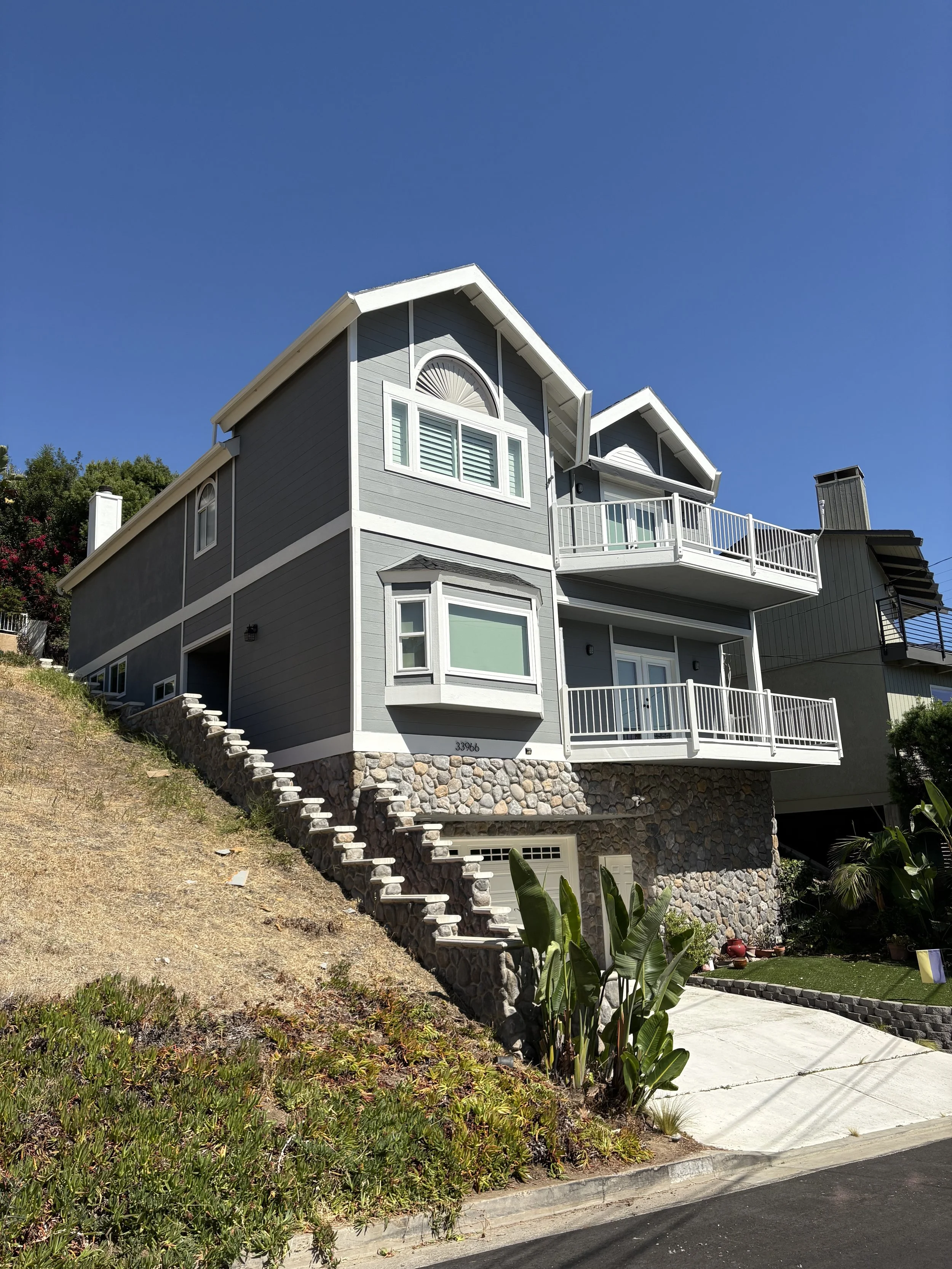 A two-story gray house with white trim, located on a hillside with stairs leading up to the entrance. The house has multiple windows and a balcony on the second floor. There is a stone foundation and some greenery around the house.