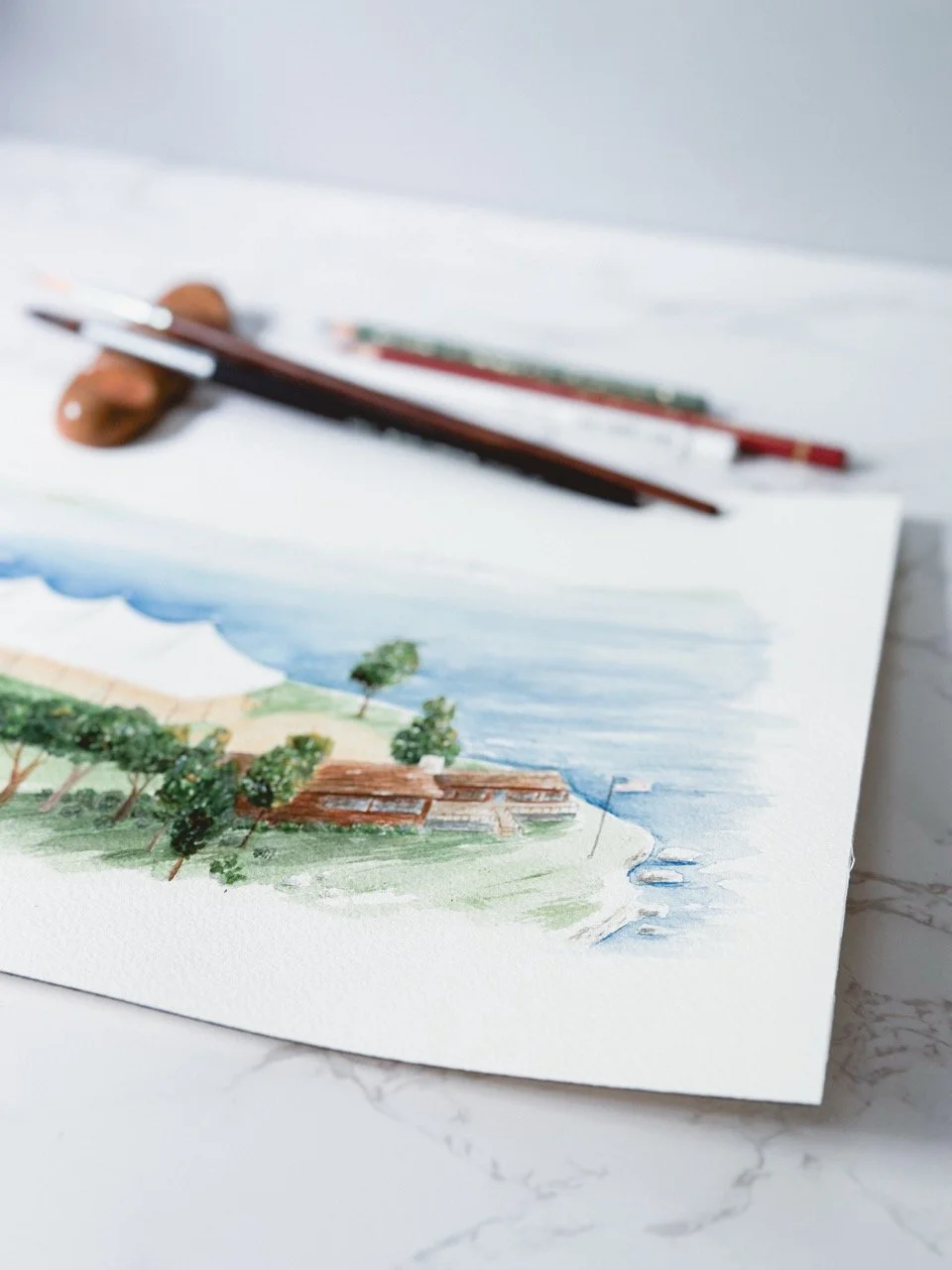 Watercolor painting of a coastal landscape with trees, a building, and the ocean, with watercolor brushes resting on top.