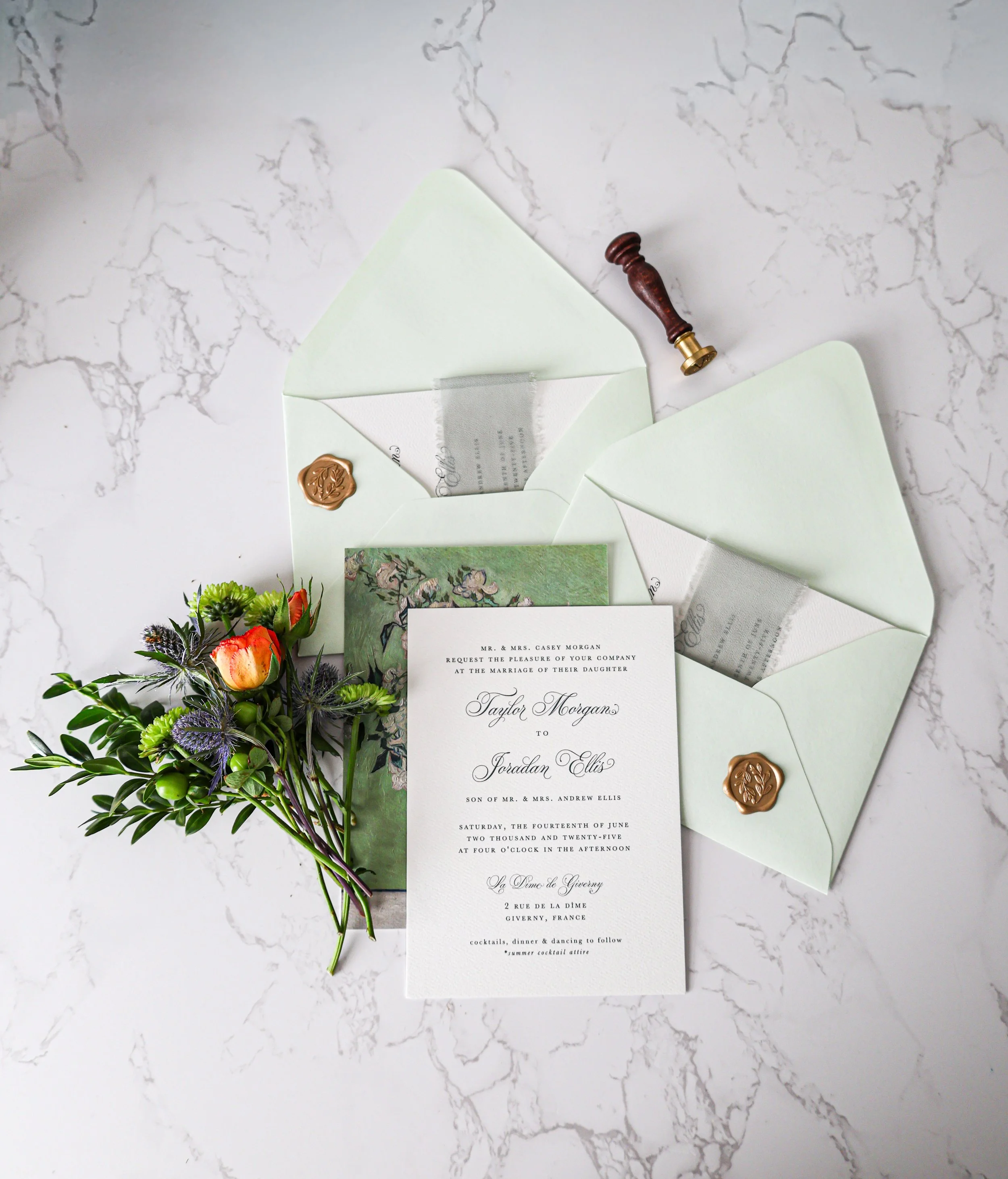 Wedding invitation with floral details, spring green envelopes with gold wax seals, a bouquet with greenery and orange flowers, and a wax seal stamp on a white marble surface.