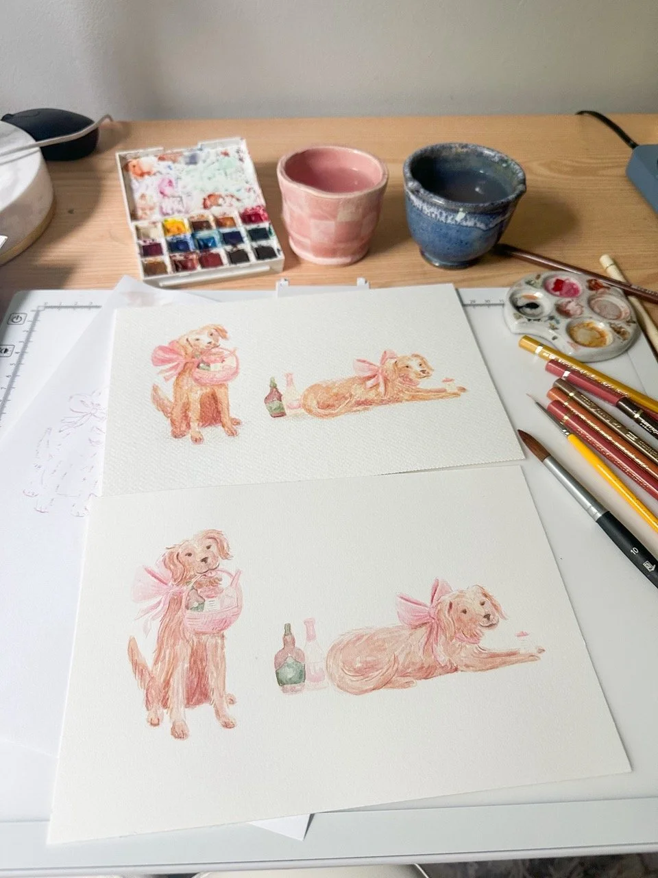 Watercolor paintings of a dog with a pink bow, holding a cake, lying down next to a bottle, displayed on a desk with watercolor supplies and art tools.