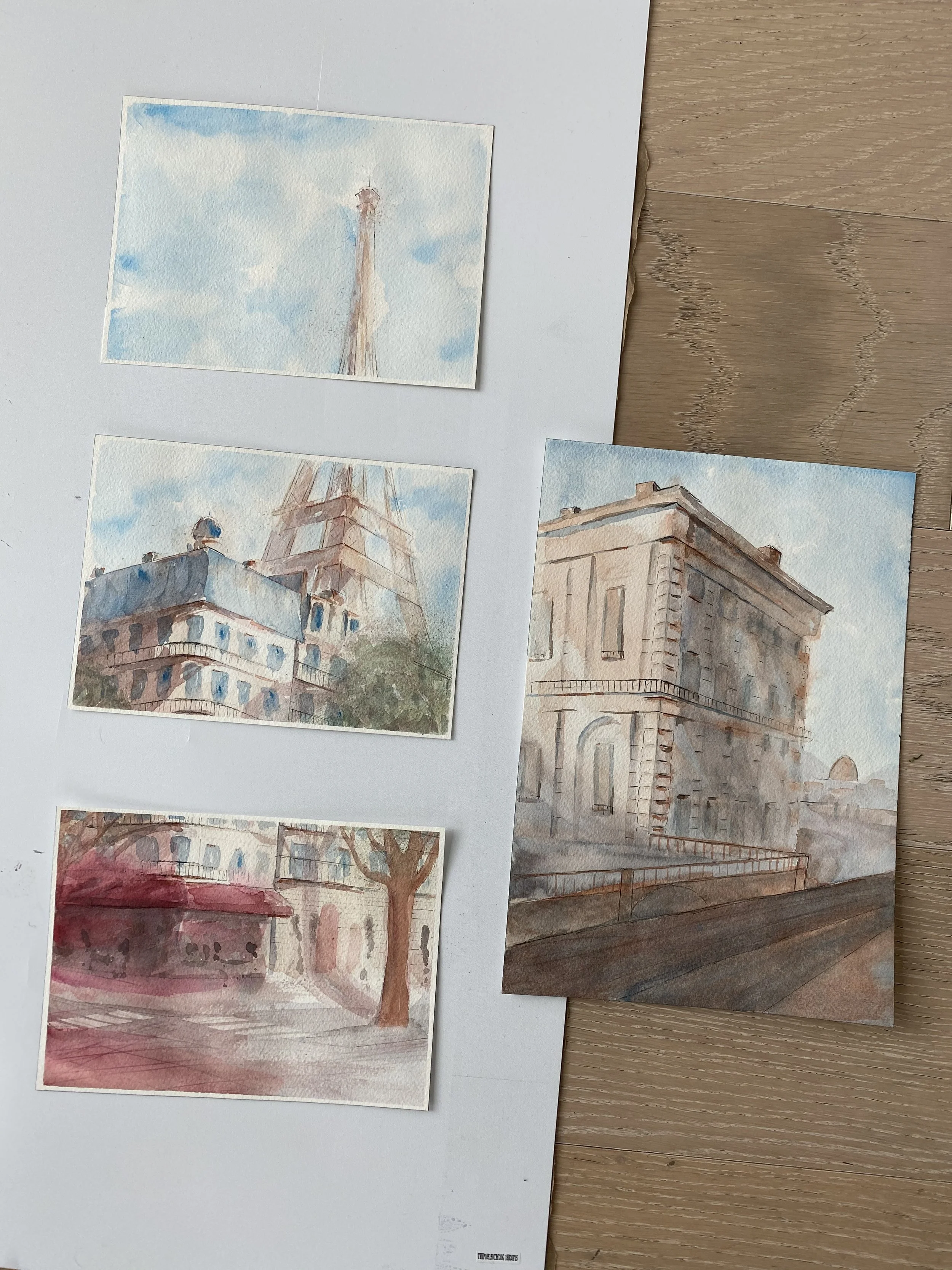 Four watercolor paintings of cityscapes and landmarks laid out on a flat surface, with one larger painting on the right showing a historic building with columns and a balcony, three smaller paintings on the left depicting a tower, a building with a s