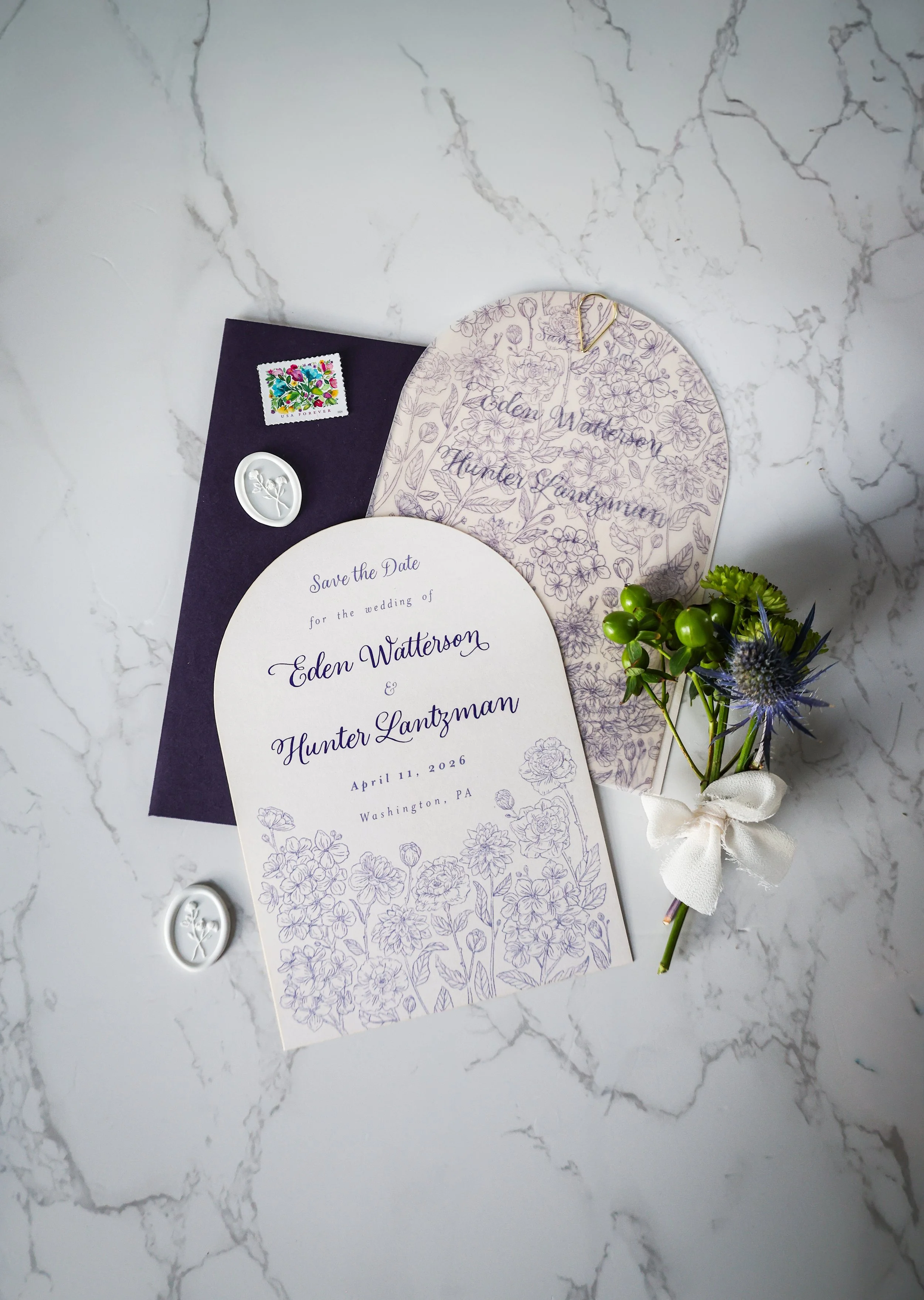 Elegant wedding save-the-date card with floral design, navy envelope with a colorful stamp, and wax seals, accompanied by a small bouquet of flowers tied with a white bow, all placed on a white marble surface.