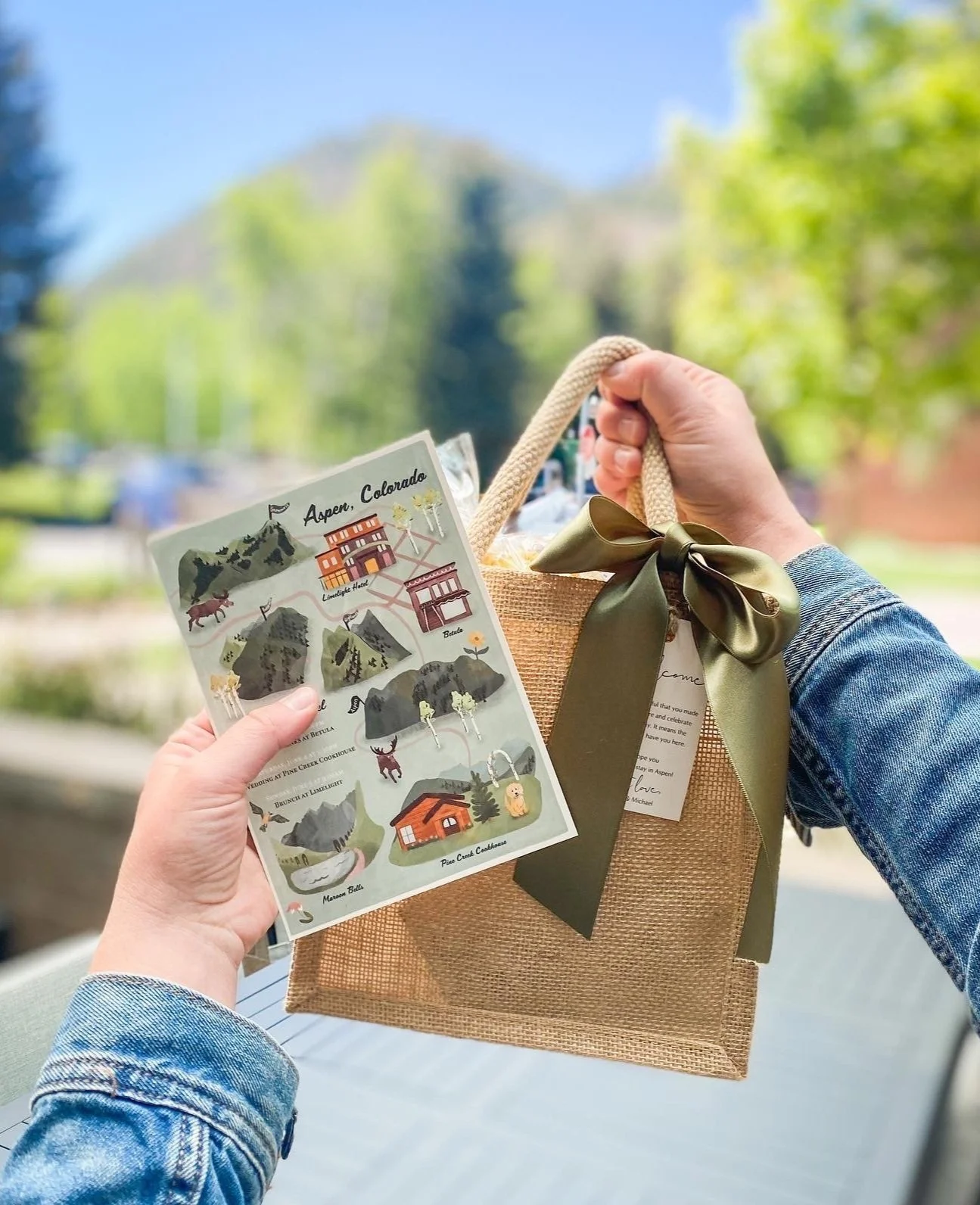 Person holding a small gift bag with a green ribbon bow and a map of Aspen, Colorado, showing local attractions, outdoors with trees and mountain in the background.