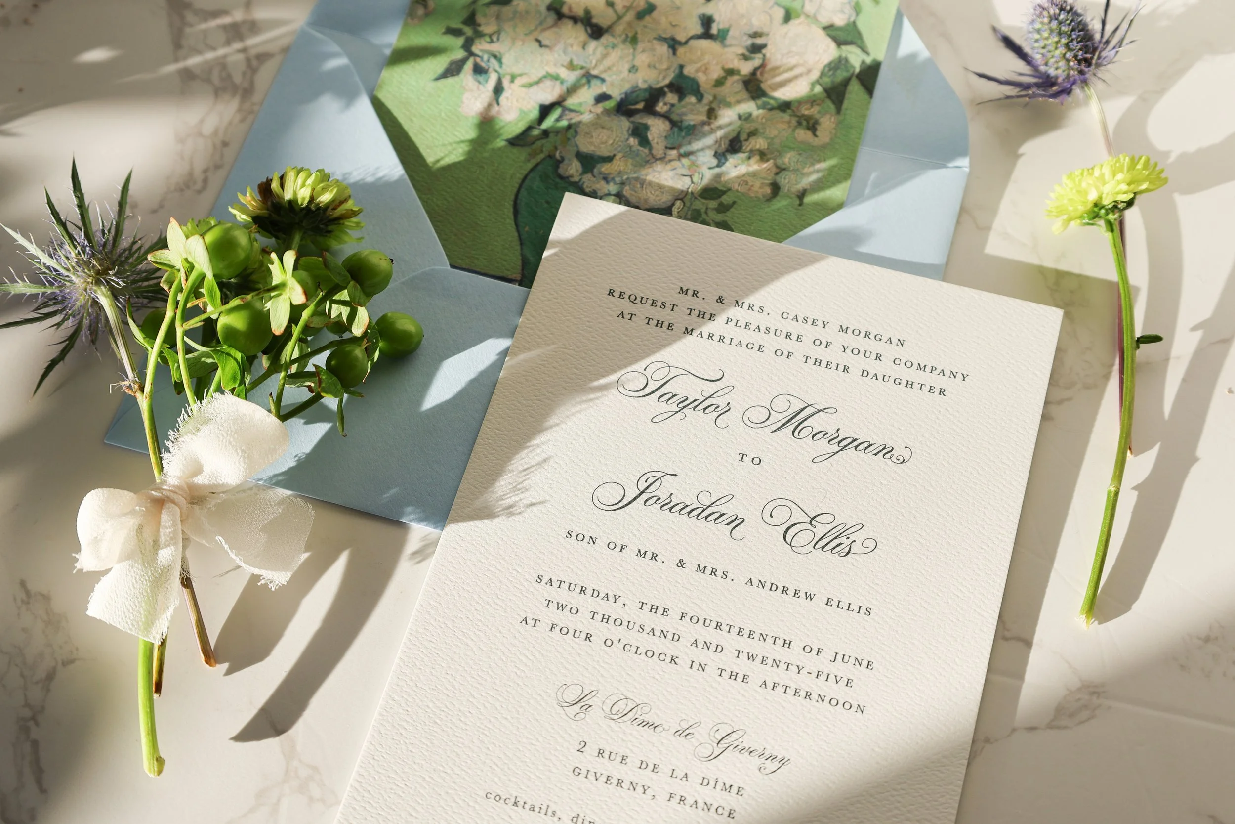 Wedding invitation with a card displaying the names Taylor Morgan and Jordann Ellis, surrounded by flowers and a blue envelope with a floral interior design.