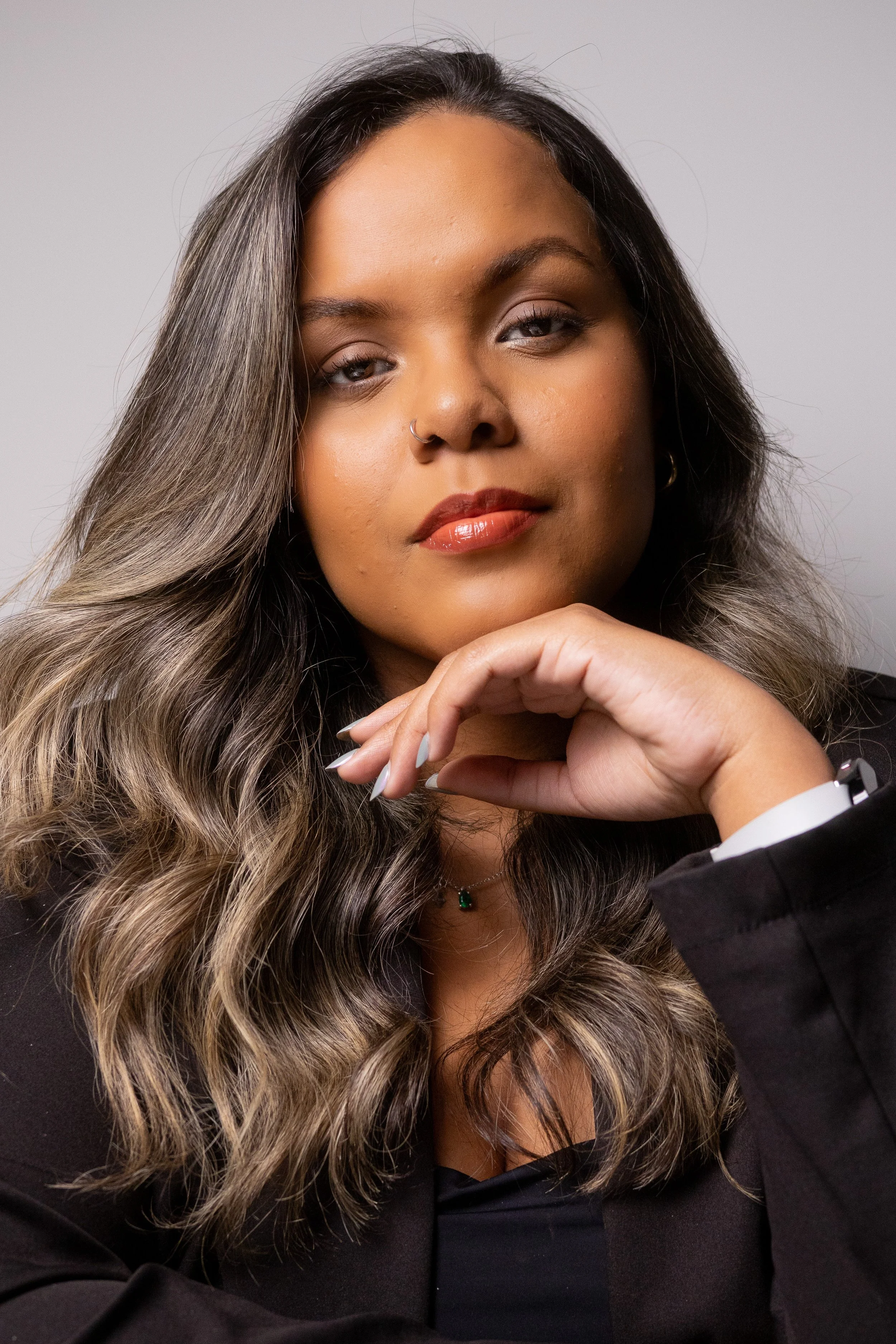 Retrato de uma mulher com cabelo castanho com luzes, maquiagem discreta, usando brinco de argola e um colar com pingente verde, vestindo blusa preta e base lisa, olhando para a câmera com expressão confiante, mãos próximas ao rosto.