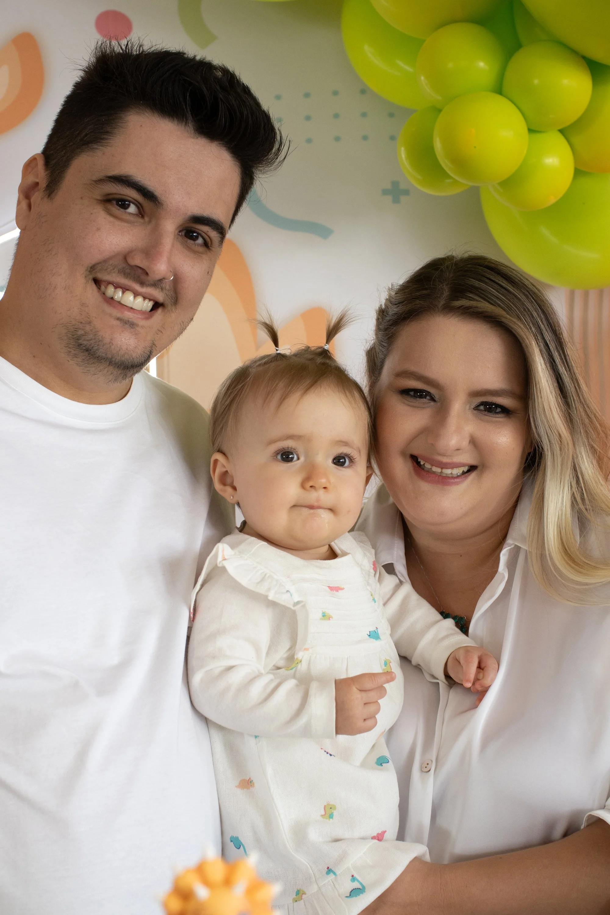 Família comemorando com balões verdes ao fundo. Homem, mulher e criança sorridentes.
