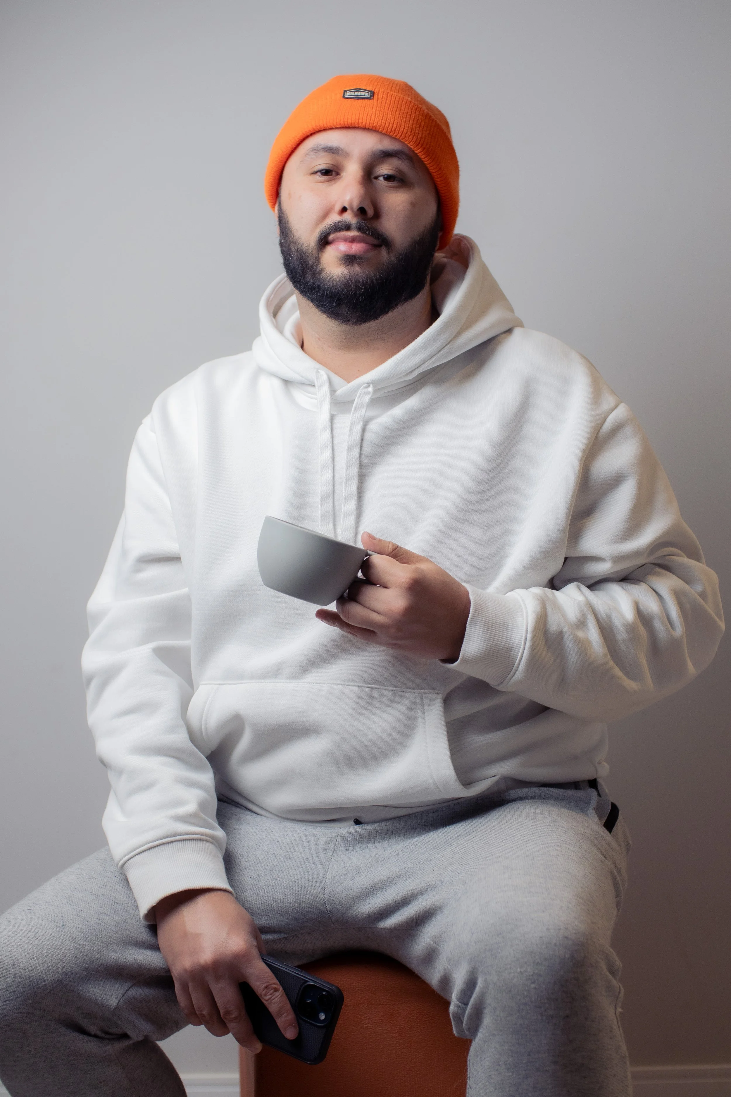 Homem com barba, vestindo moletom branco e gorro laranja, sentado segurando uma xícara e um celular
