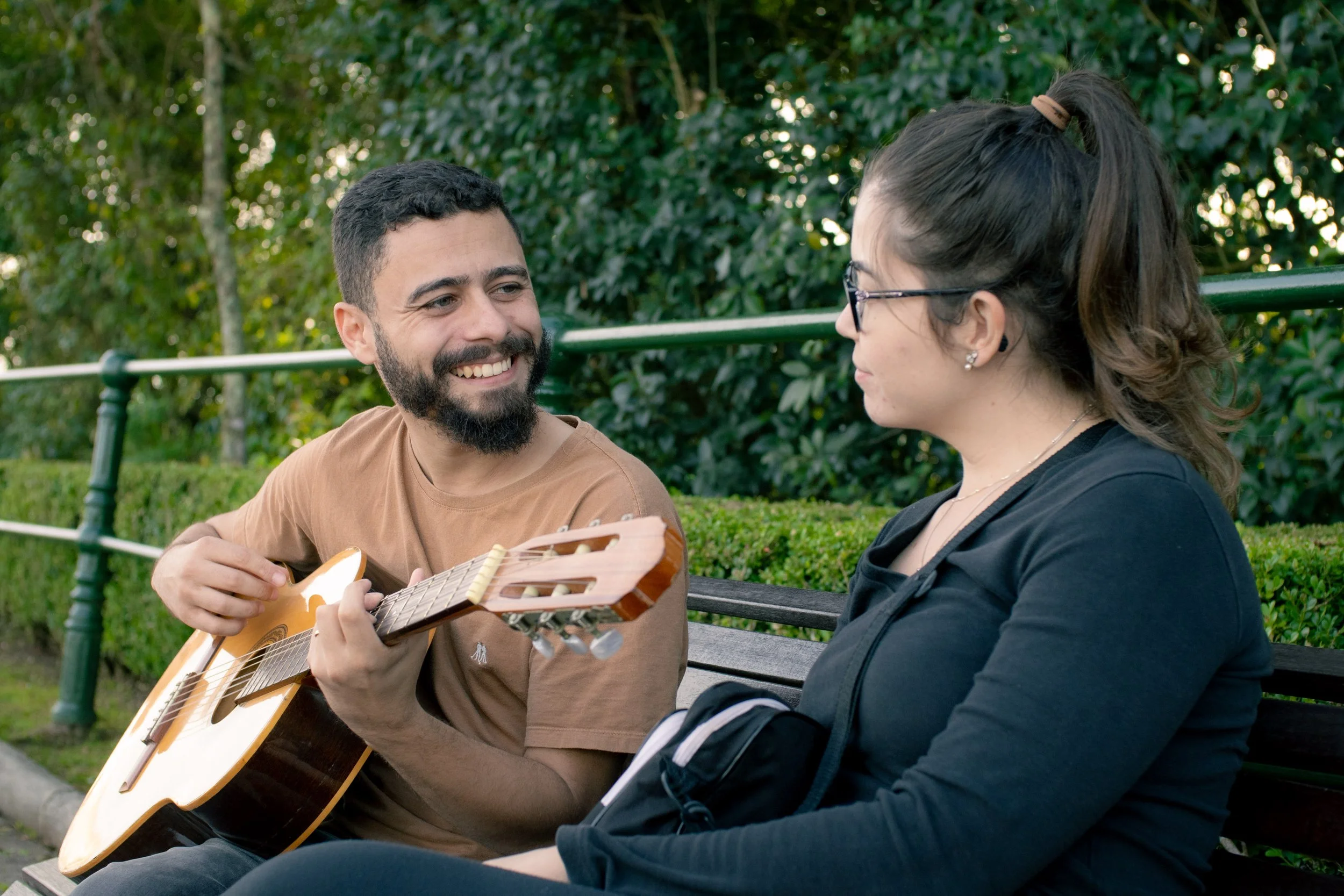 Homem tocando violão e mulher ouvindo enquanto sentam em banco de parque cercado por vegetação.
