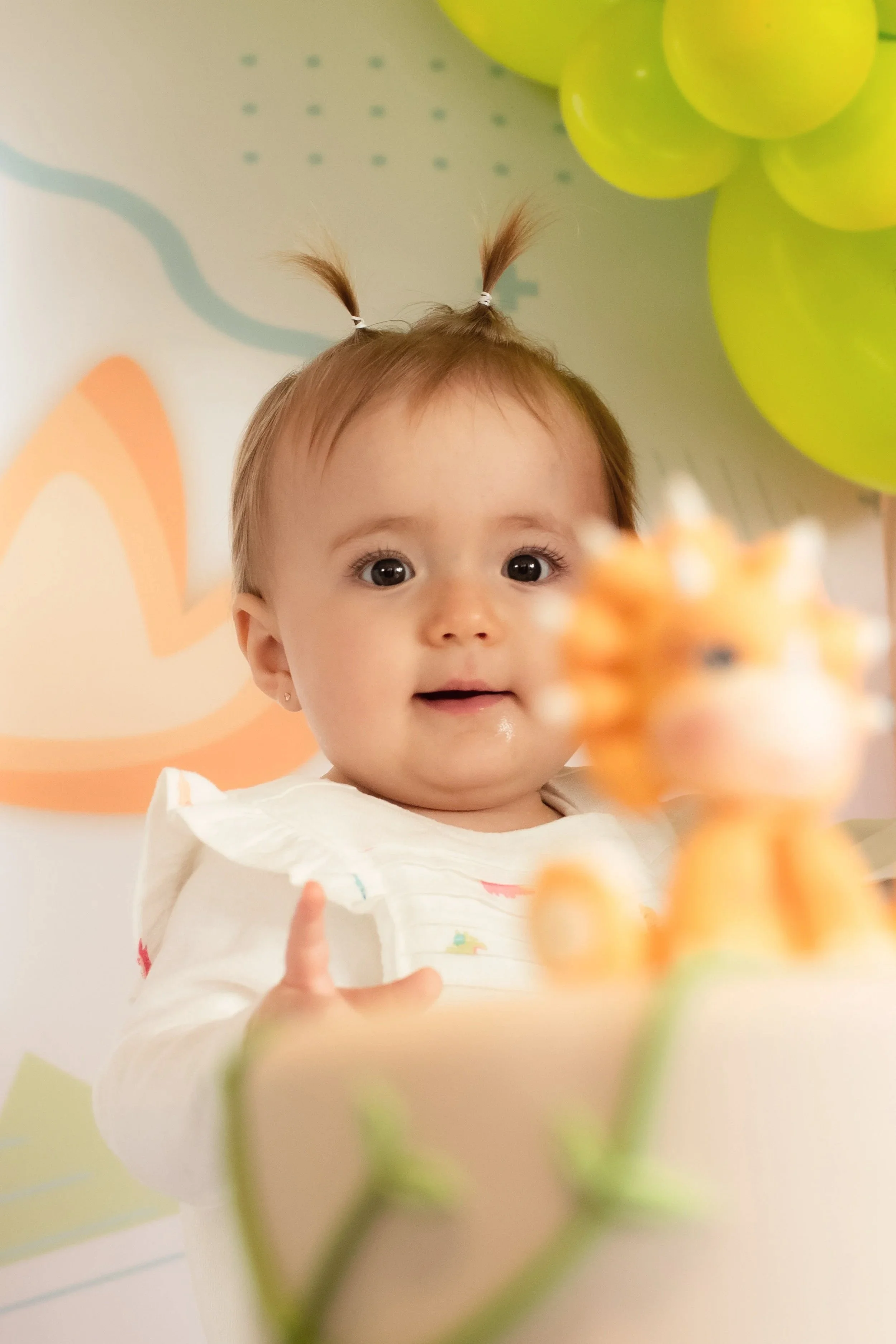 Bebê com cabelo preso em duas pontas, olhando para uma figura de um leão de pelúcia, com balões verdes ao fundo.