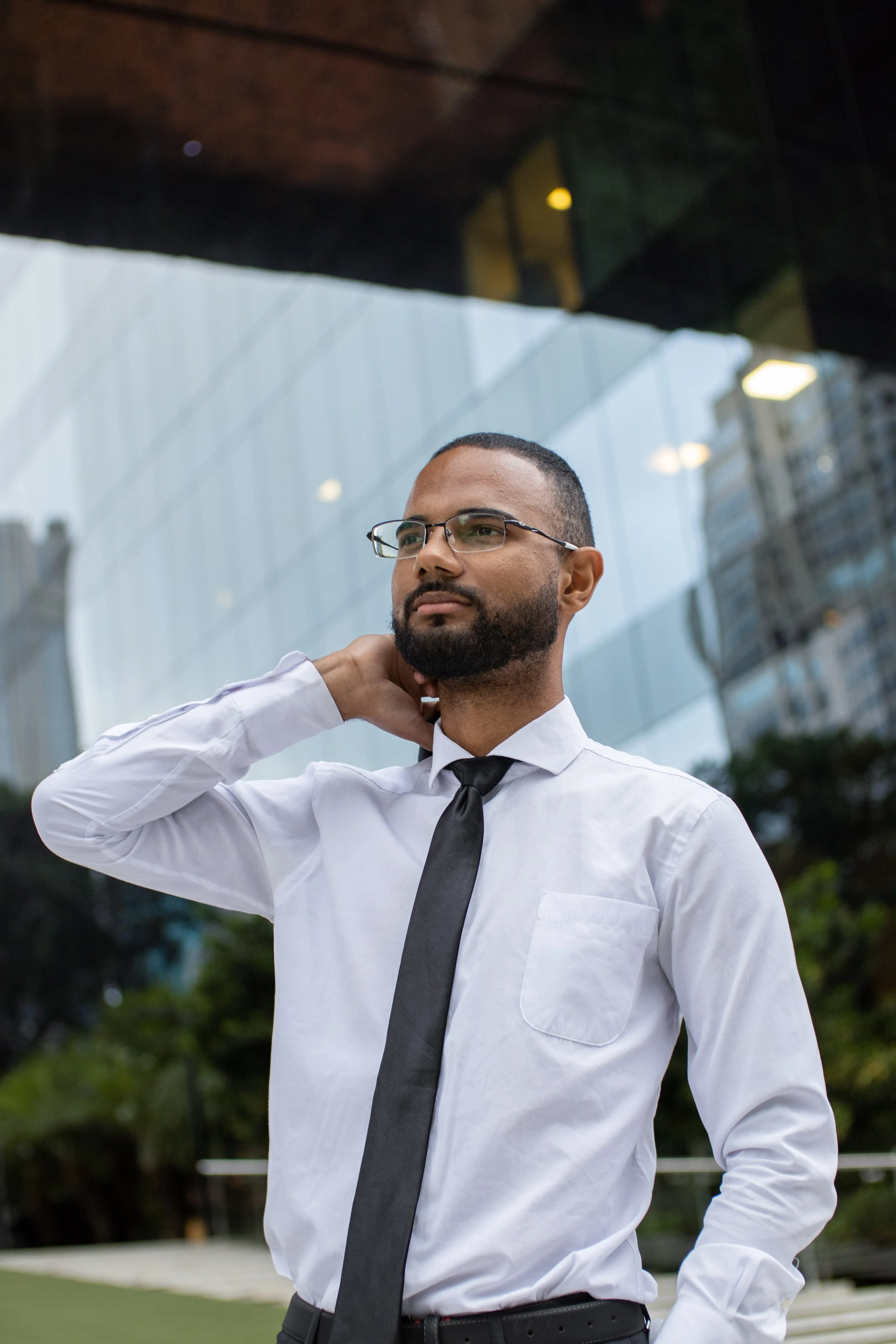 Homem de terno branco, gravata preta e óculos, com barba e cabelo curto, em ambiente urbano ao ar livre, com reflexo de prédios de vidro ao fundo.