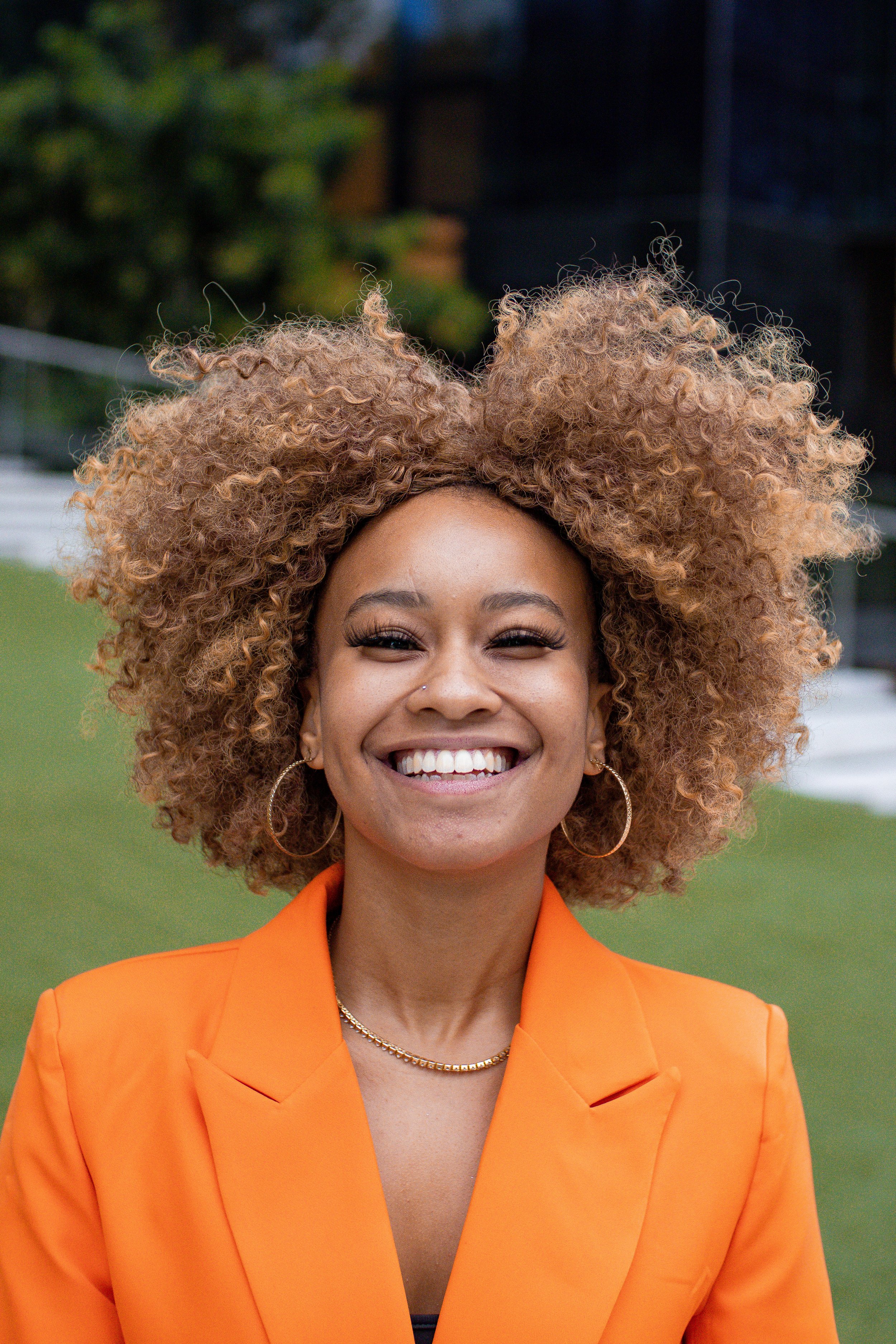Mulher sorrindo ao ar livre, com cabelo cacheado e vestindo blusa laranja e acessórios dourados.