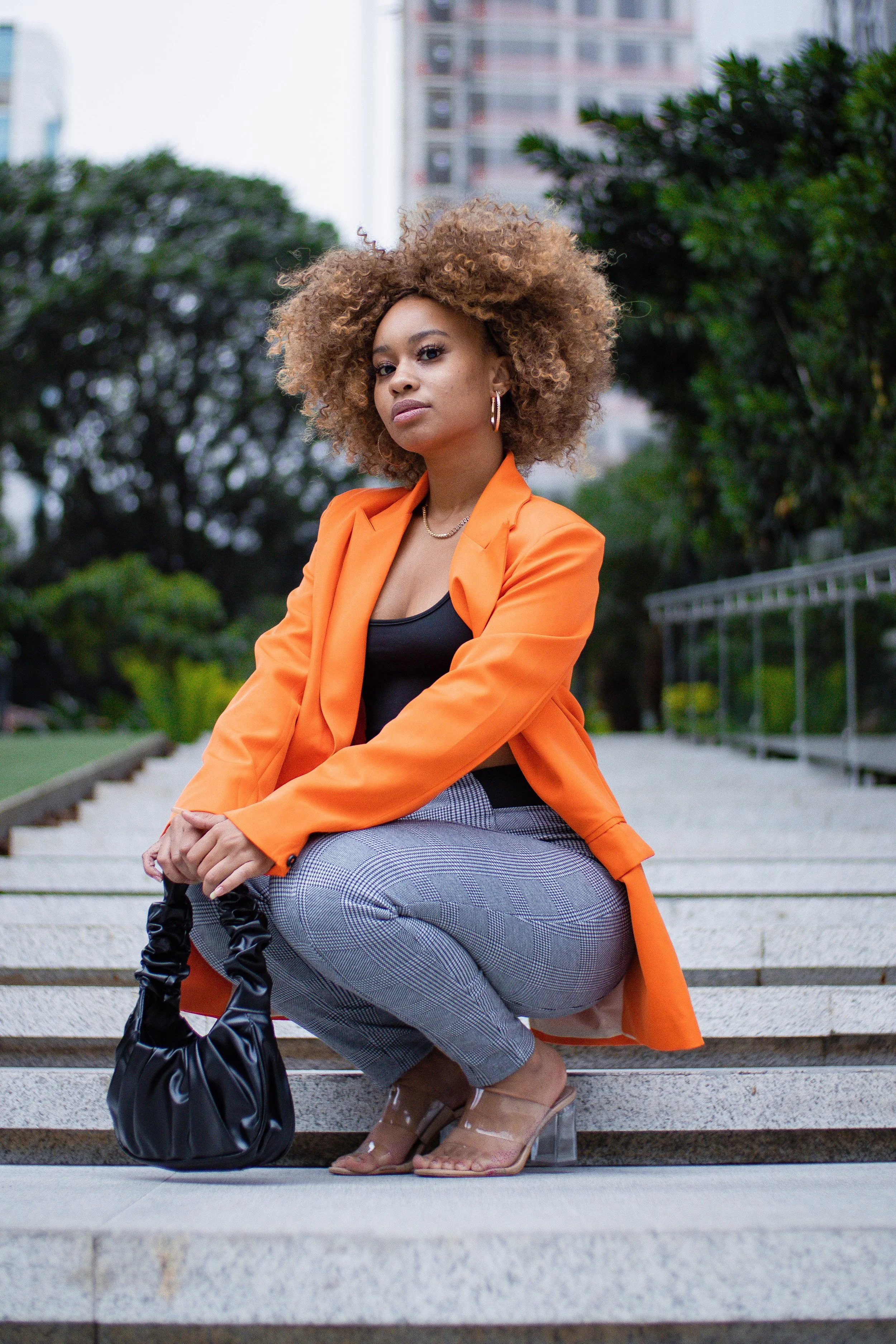 Mulher com cabelo cacheado, usando blusa preta e blazer laranja, calça de padrão xadrez e sapatos de salto, segurando uma bolsa preta, posando em escadaria ao ar livre com árvores e edifícios ao fundo.