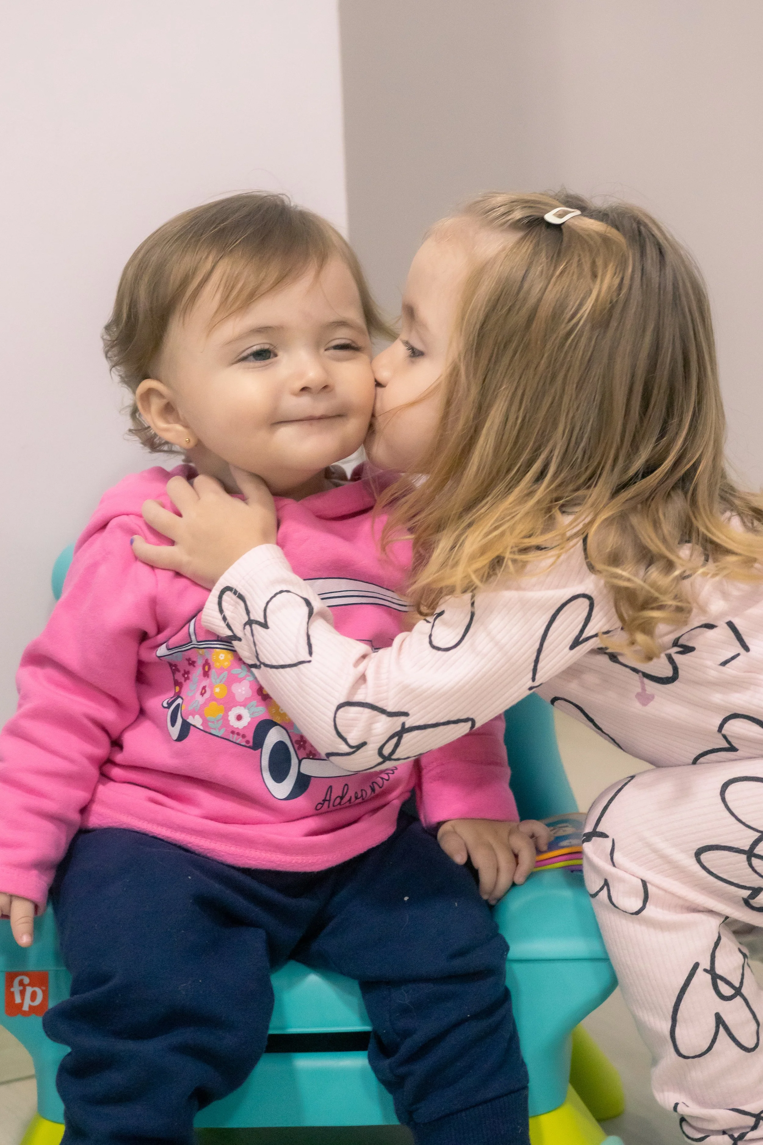 Duas crianças, uma menina mais velha beijando carinhosamente uma menina mais nova na bochecha, enquanto ela está sentada em uma cadeira de plástico azul