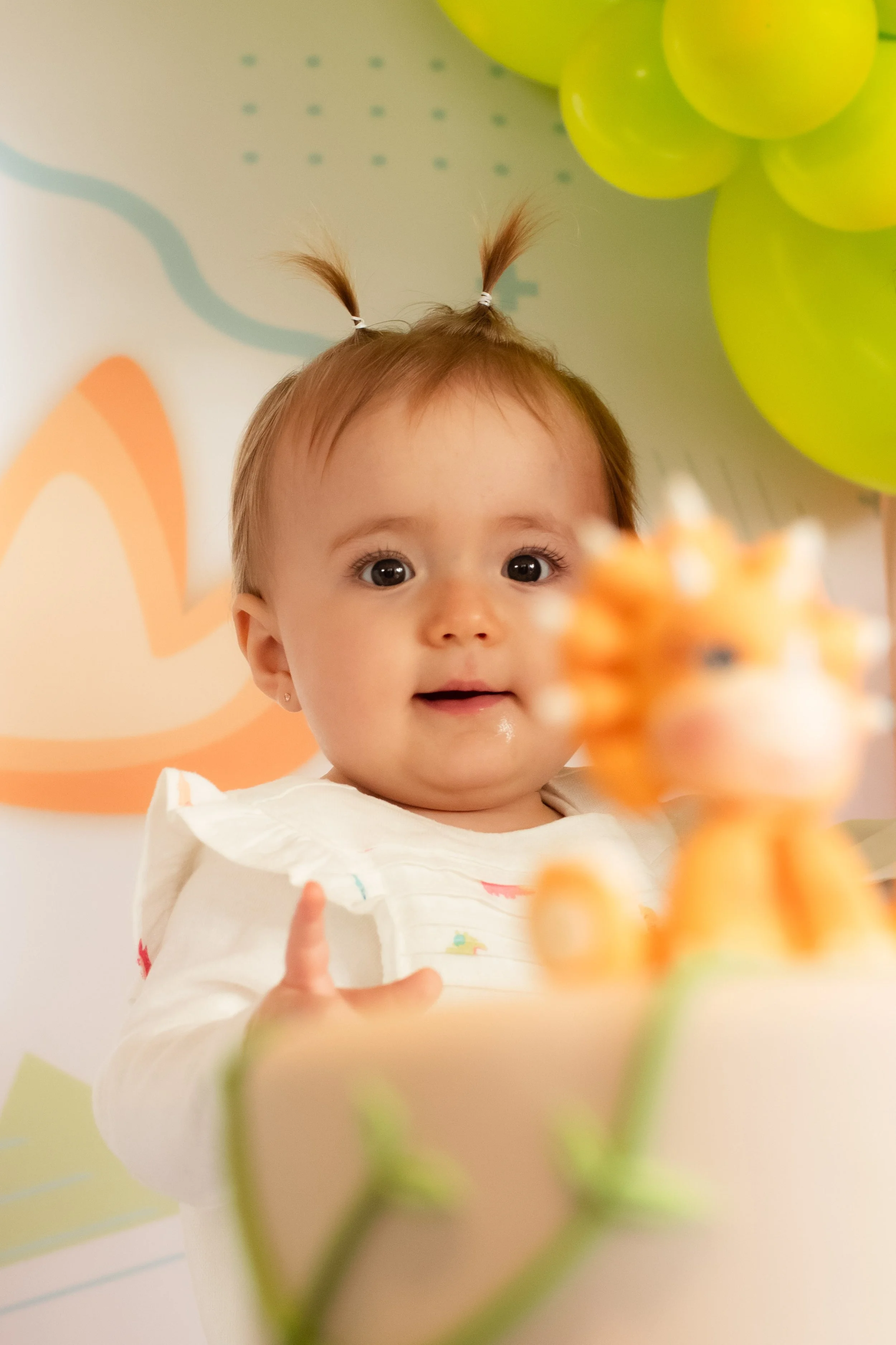 Bebê com cabelinho escuro e duas pequenas pontas amarradas, olhando para uma vela de aniversário com uma expressão de surpresa ou encanto, ao fundo balões verdes e decoração de festa infantil.