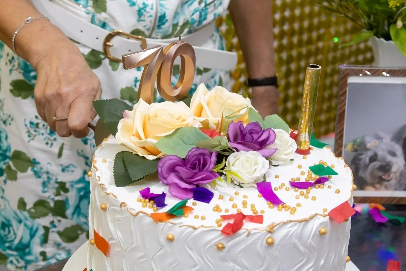 Bolo de aniversário decorado com flores artificiais e confetes coloridos, com número 70 dourado como topper, e uma vela dourada acesa ao lado.