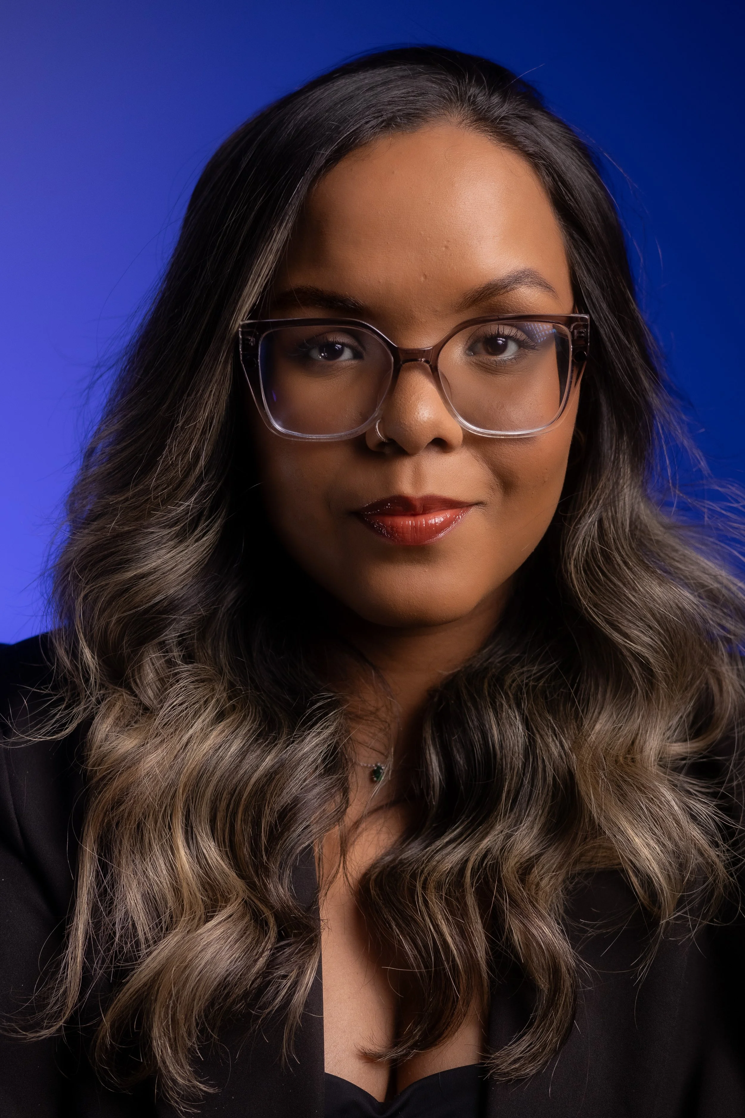 Retrato de uma mulher com cabelos longos e ondulados, óculos grandes, batom escuro e roupa preta, contra fundo azul.