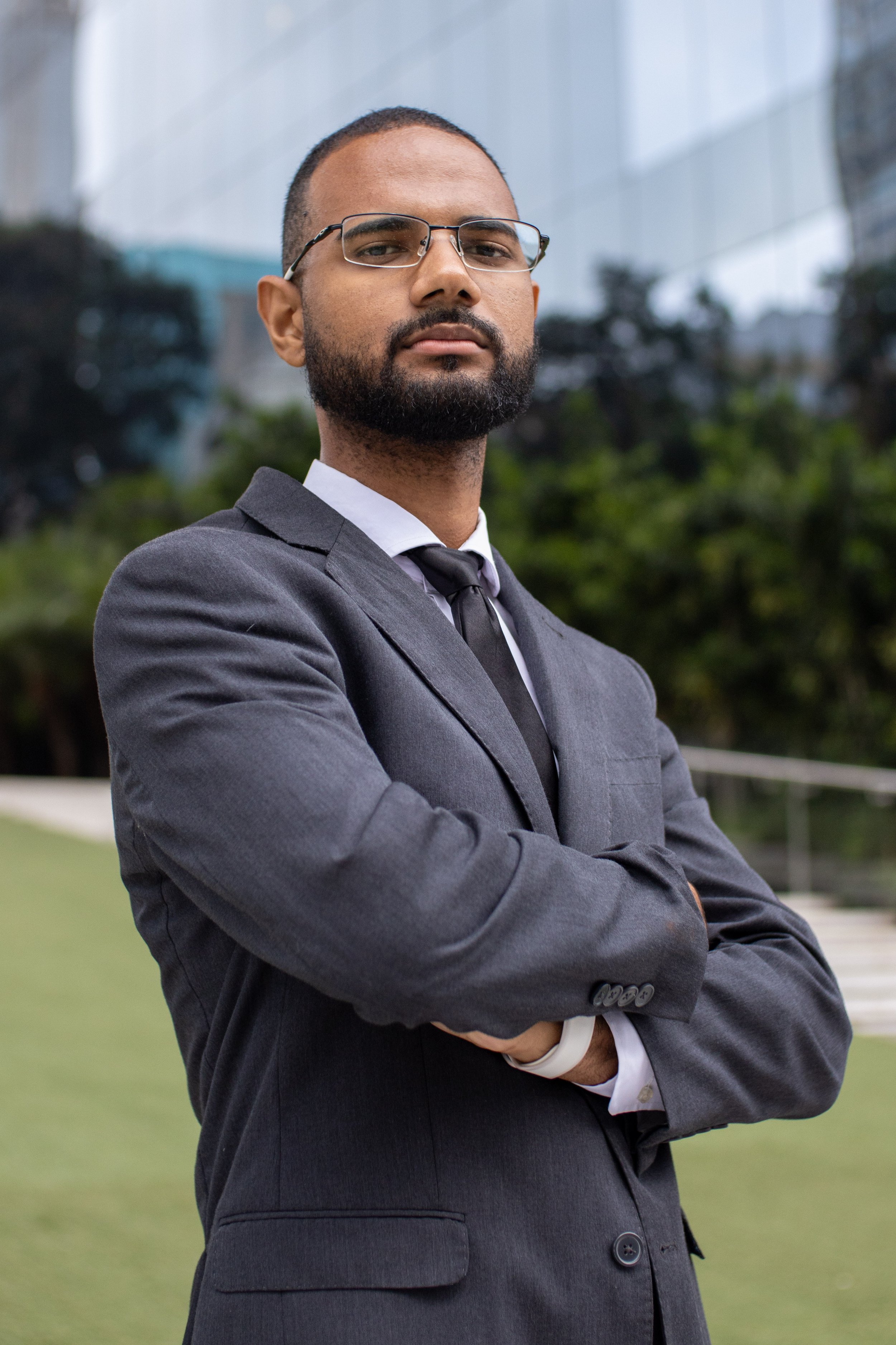 Homem de terno cinza, com braços cruzados, usando óculos e gravata, em ambiente externo com prédios ao fundo.