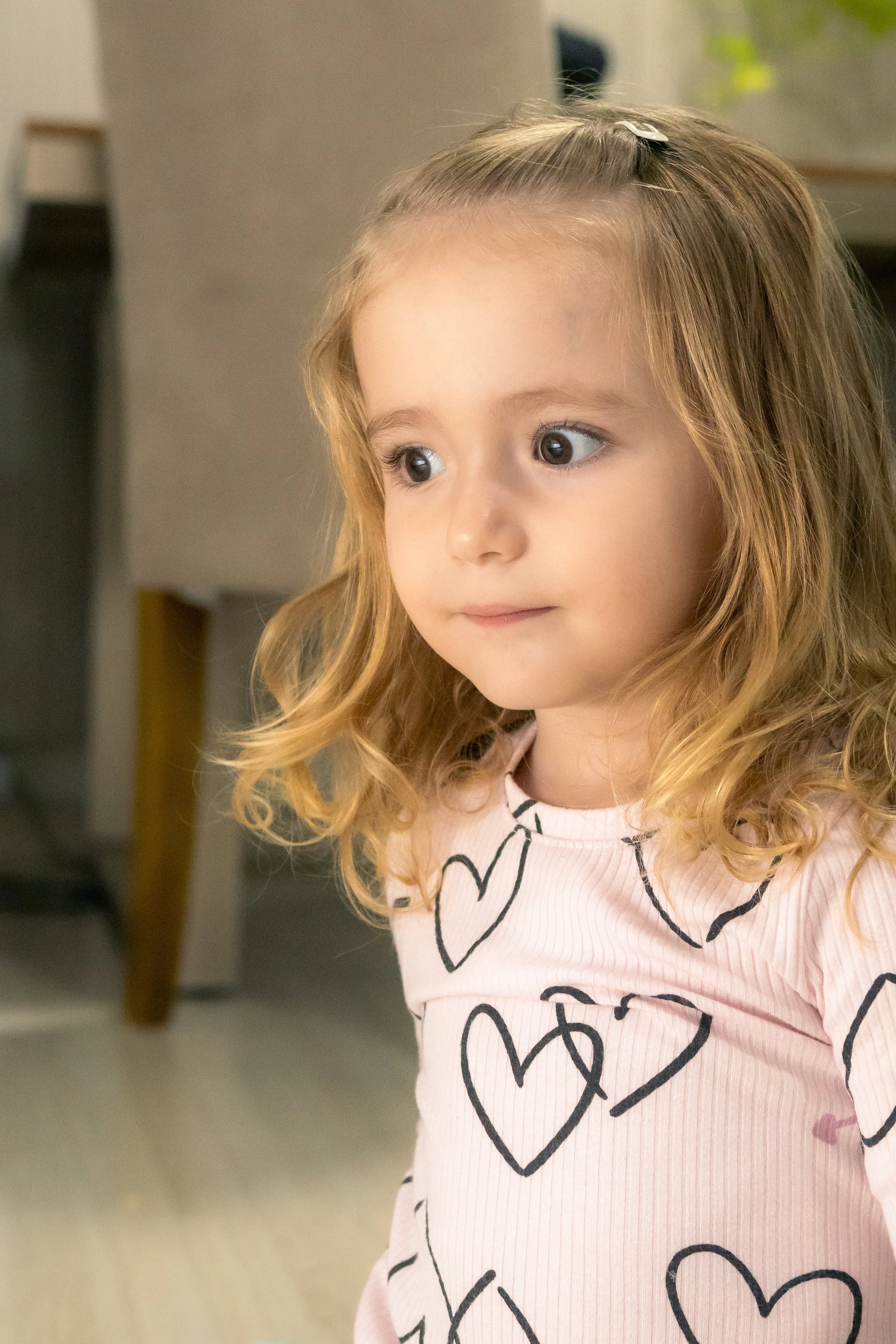 Menina de cabelos cacheados e longos, usando blusa rosa com corações pretos, olhando para o lado, em ambiente interno.