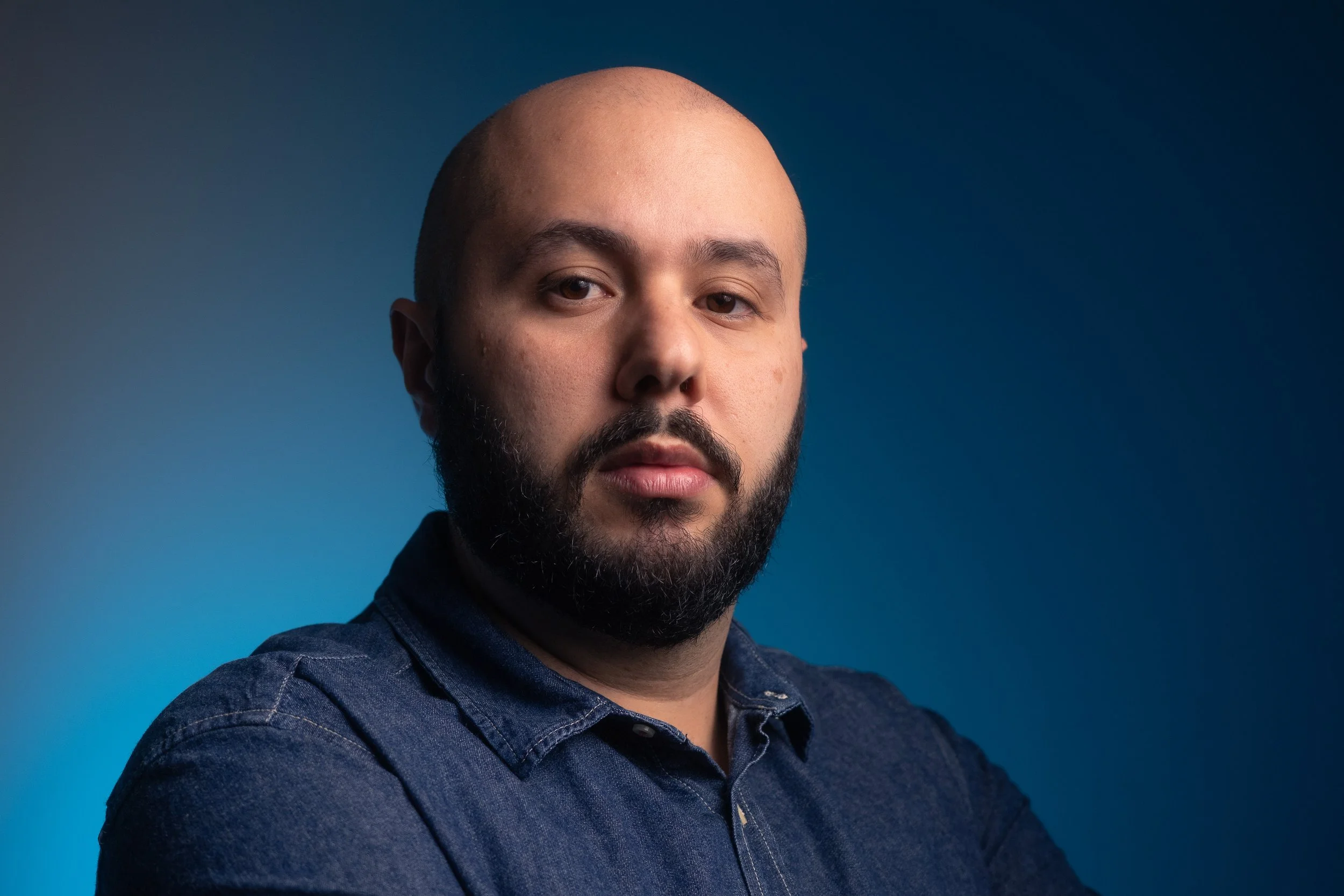 Retrato de um homem com barba, usando uma camisa azul escura, contra um fundo azul gradiente.