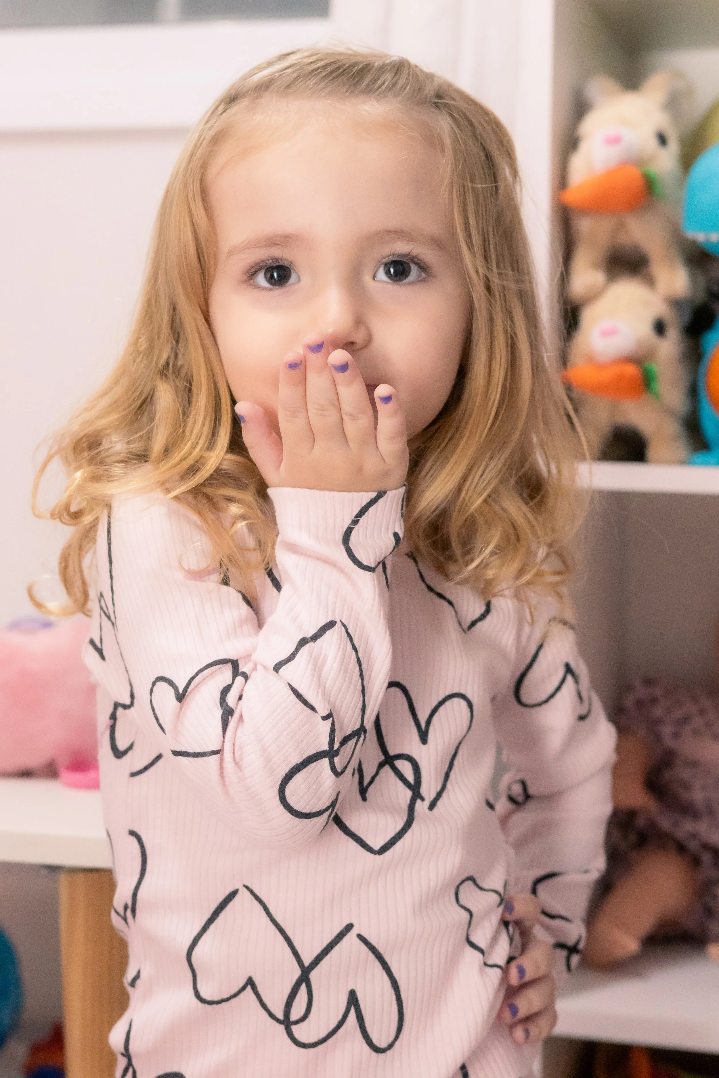 Menina com cabelo cacheado, roupa rosa com corações pretos, cobrindo a boca com a mão, em um ambiente de brinquedos.