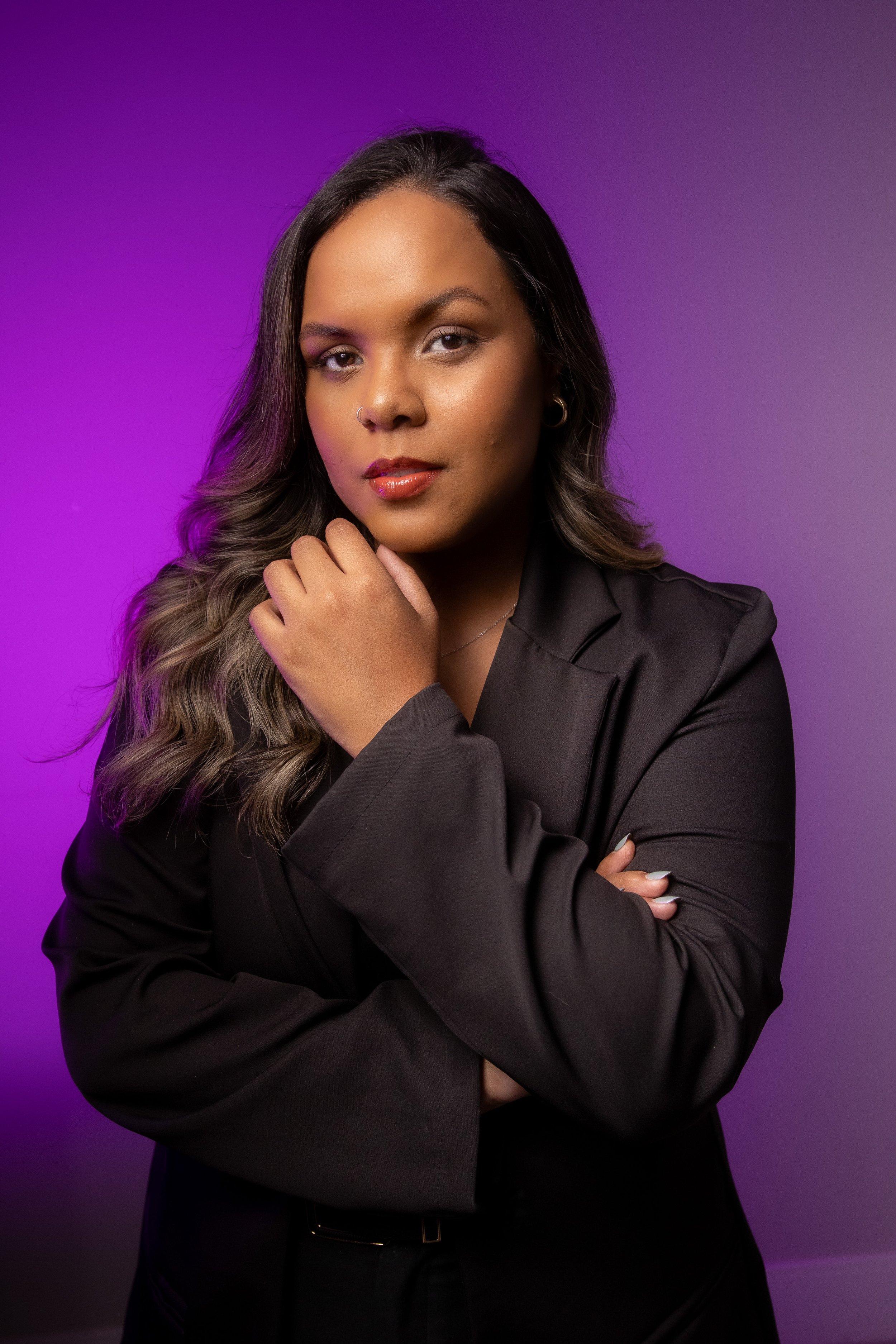 Mulher com cabelo ondulado e pele morena, usando blazer preto, posando com a mão no queixo, fundo roxo com degradê.