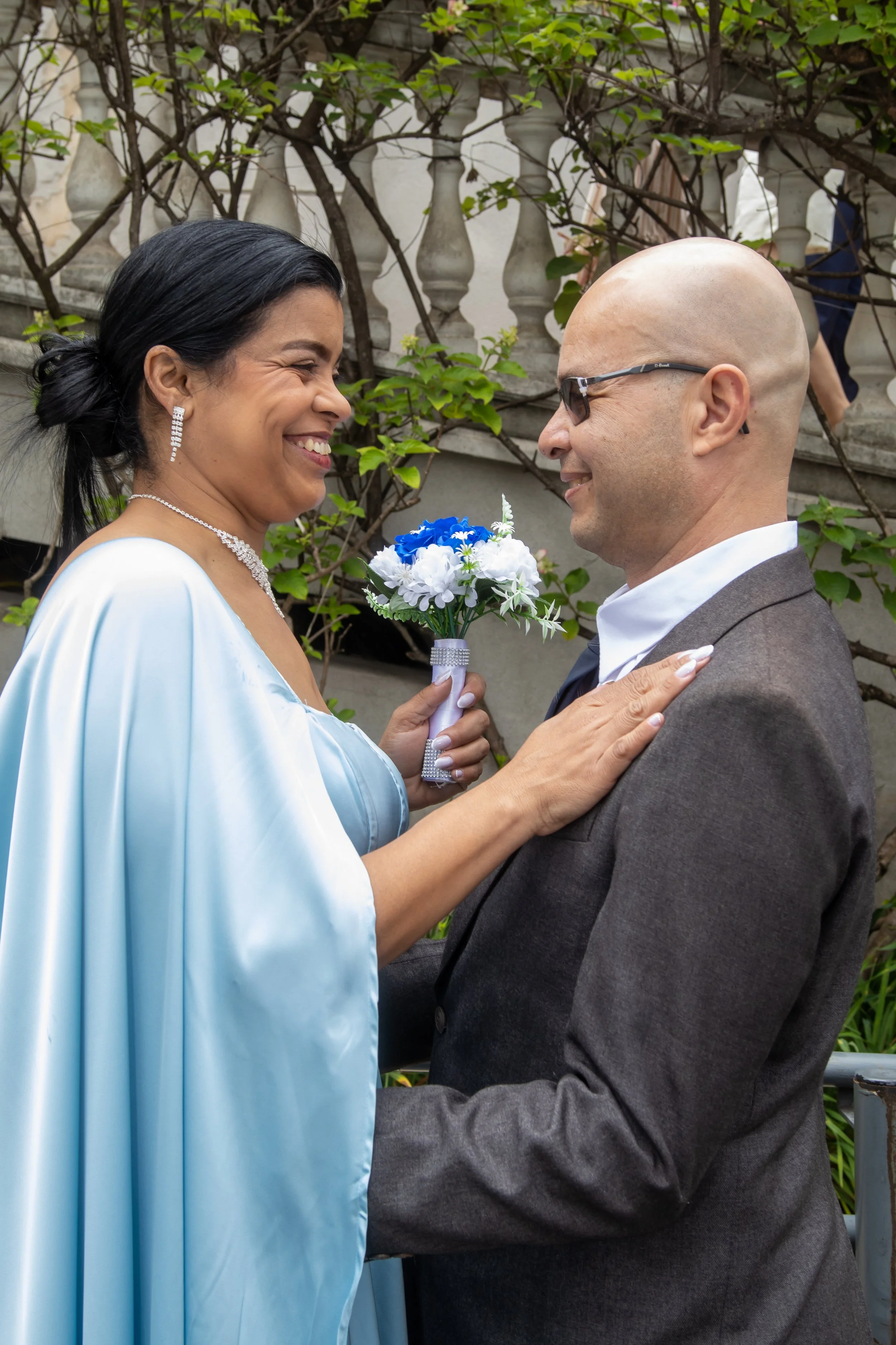 Casal sorrindo em uma cerimônia de casamento ao ar livre, com mulher vestindo vestido azul e homem de terno, segurando um buquê de flores azuis e brancas.