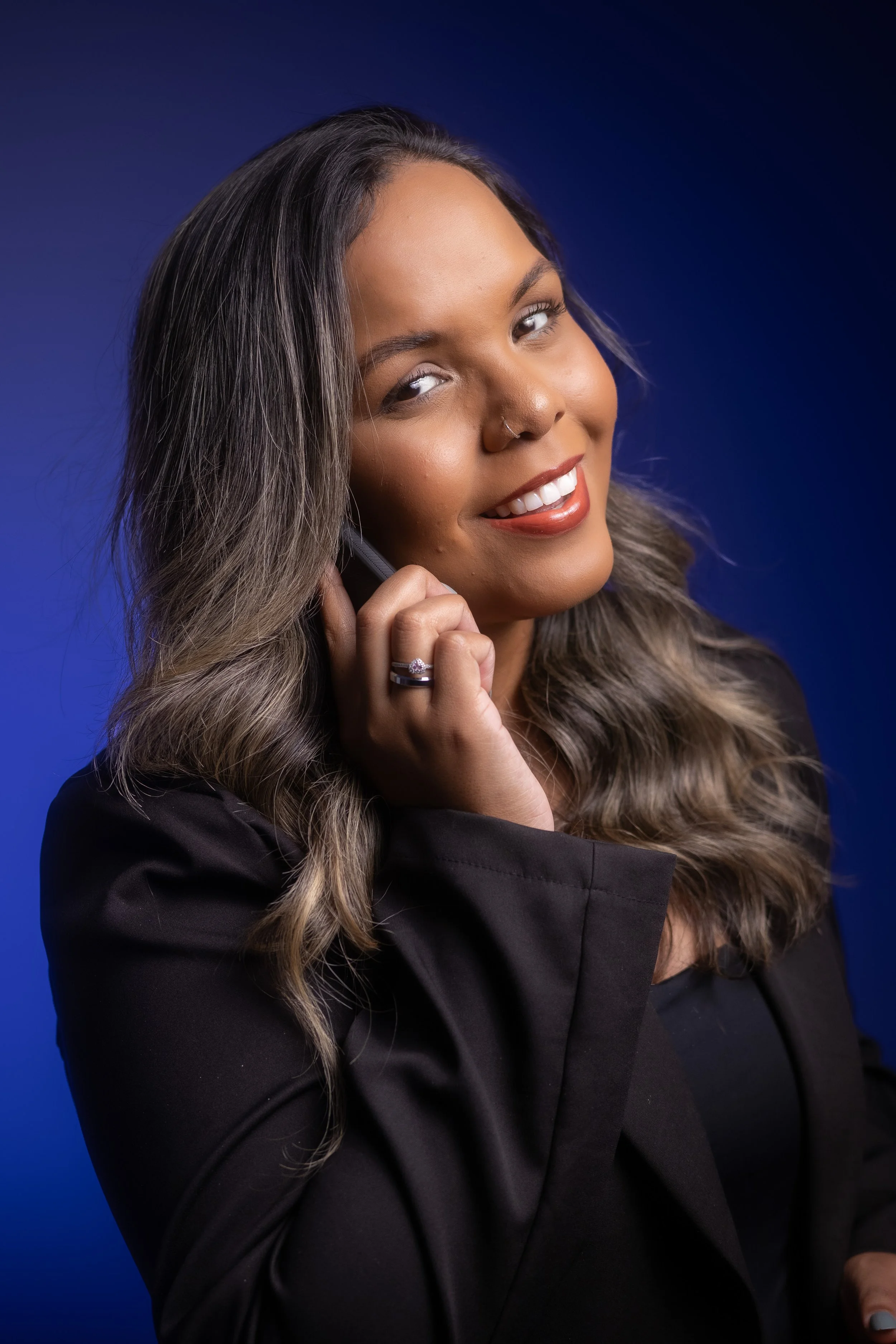 Mulher sorridente usando blazer preto, com cabelo ondulado, falando ao telefone contra fundo azul.