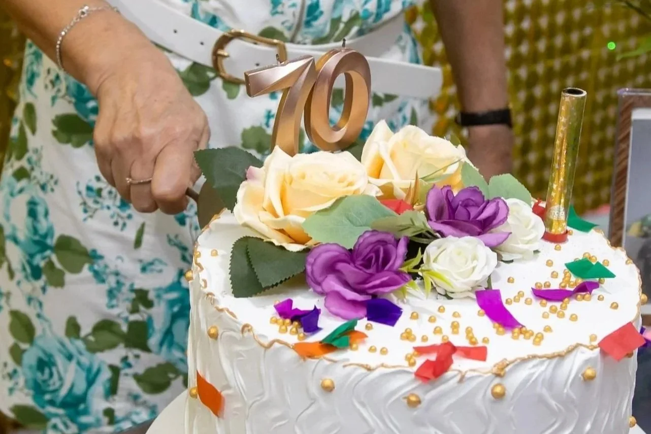 Pessoa segurando uma faca decorada cortando um bolo de aniversário branco decorado com flores artificiais e confetes coloridos, com um número dourado '70' como topo