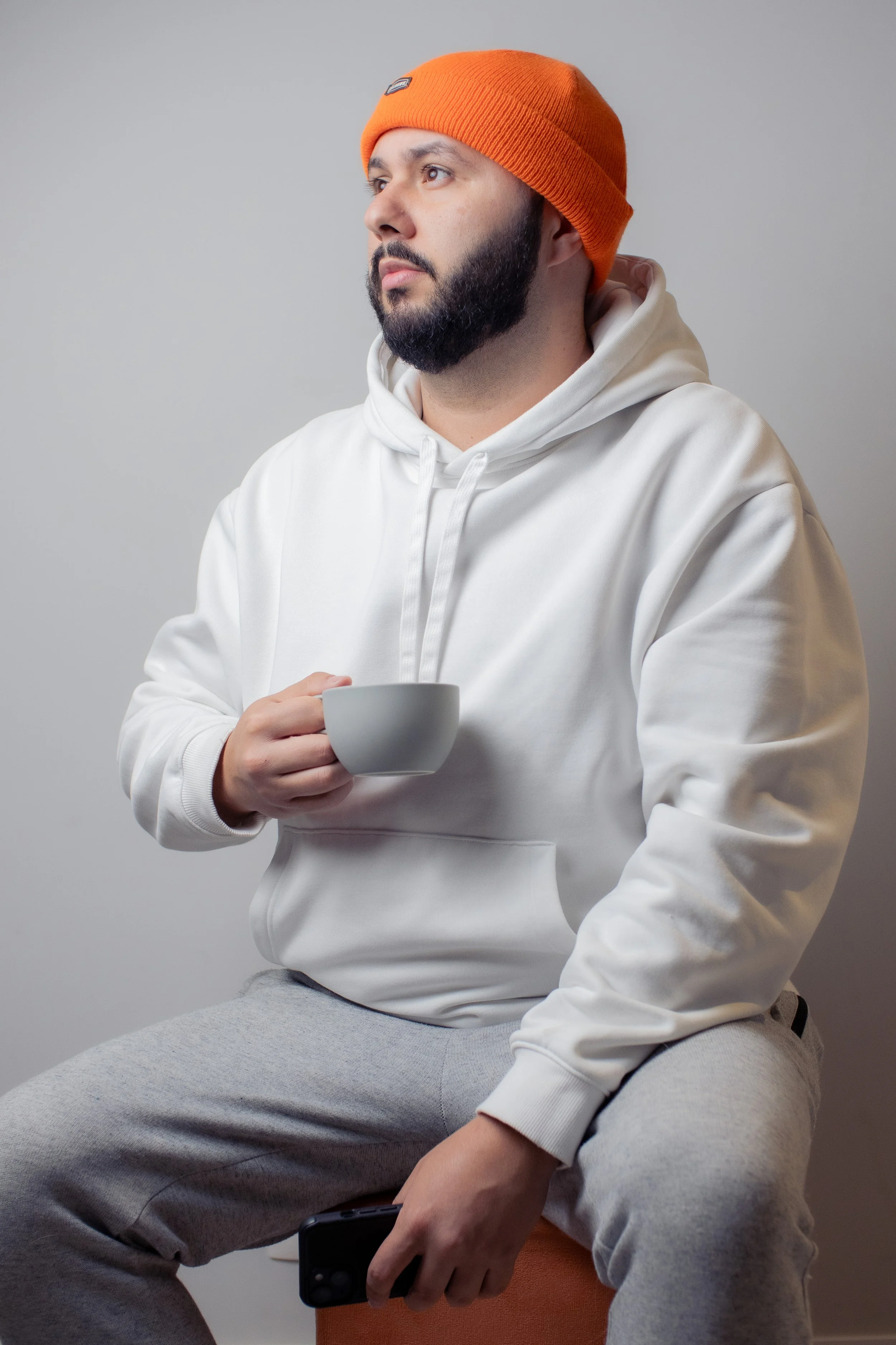 Homem de cabelo tingido de azul, barba, usando suéter branco e gorro laranja, sentado segurando uma xícara branca e um celular em um fundo cinza.