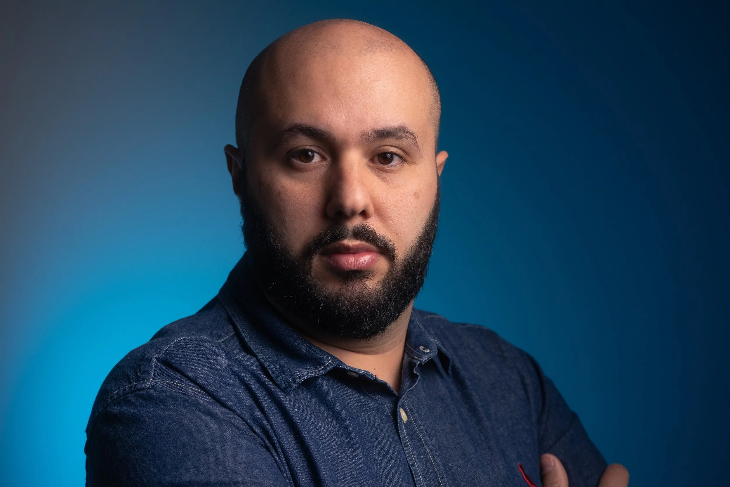 Retrato de um homem de pele clara, barba e cabeça raspada, usando camisa azul escura, cruzando os braços, contra fundo azul.